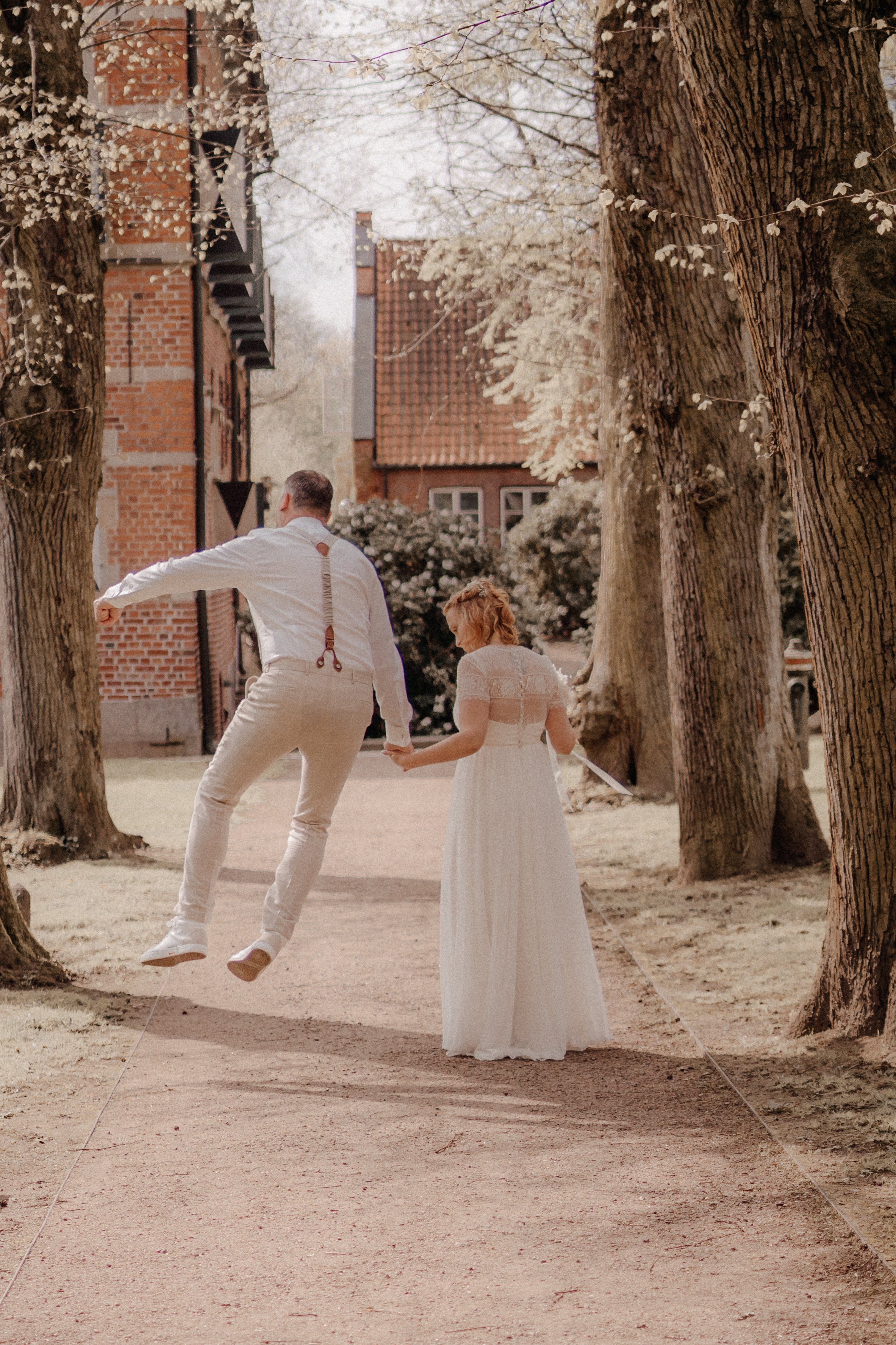 Ein Brautpaar hält Händchen auf einer Allee mit großen Bäumen und historischen Gebäuden im Hintergrund, die Szene ist sommerlich und romantisch.