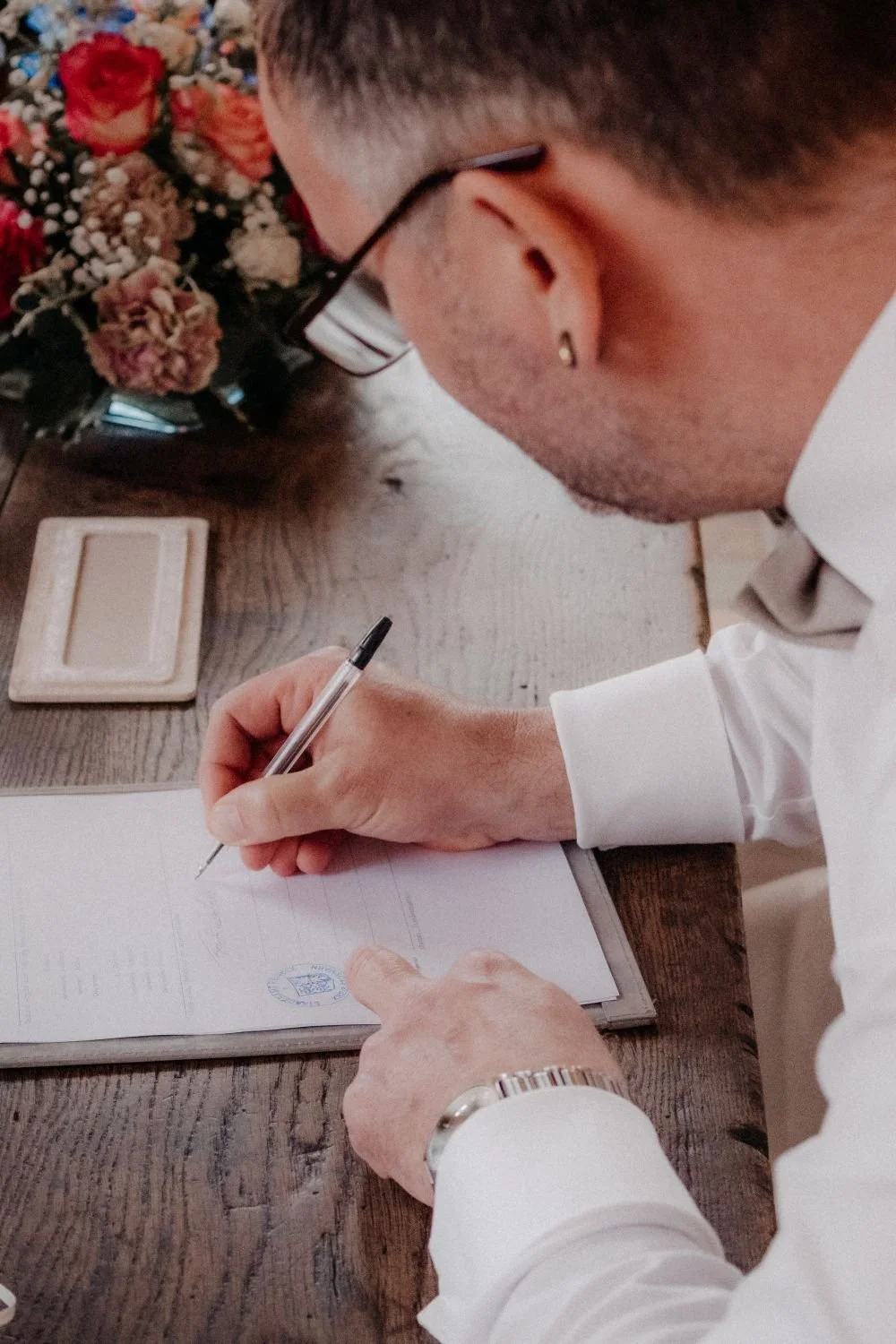 Ein Mann, der eine Unterschrift auf einem Dokument leistet, sitzt an einem Holztisch neben einem Blumenstrauß und einem Stempelkissen.