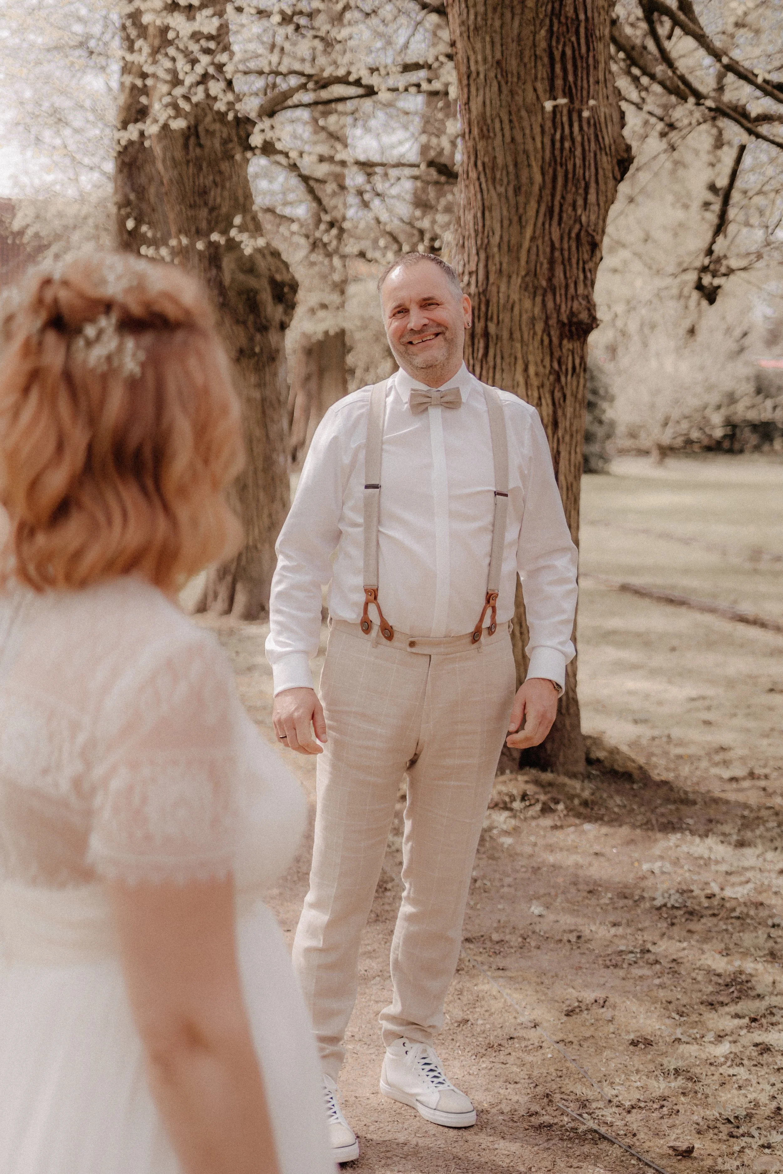 Ein Mann in formeller Kleidung mit Fliege und Hosenträgern steht vor einem Baum im Freien, lächelnd, bei Tageslicht, wahrscheinlich bei einer Hochzeit oder einem besonderen Anlass.