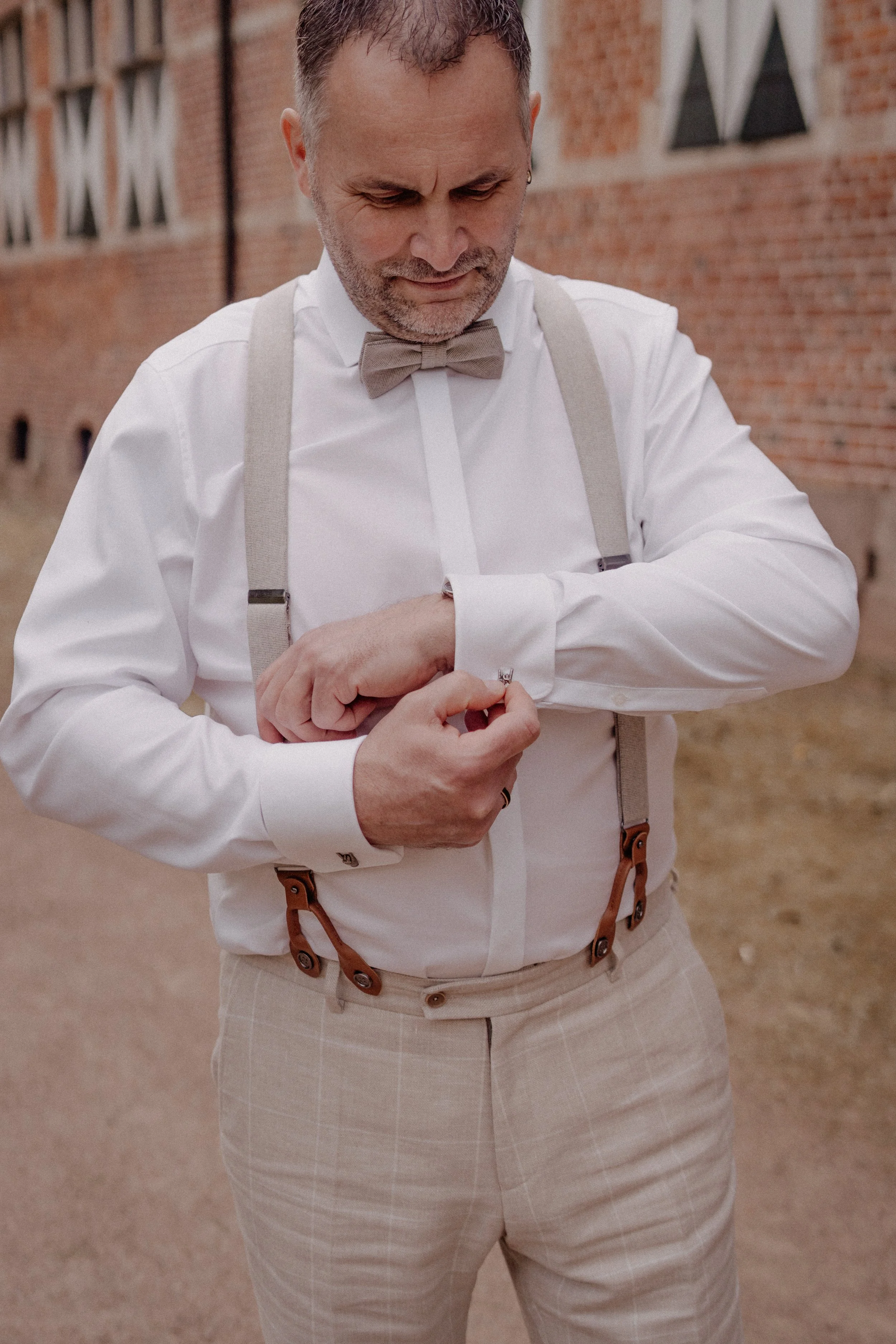 Ein Mann in formeller Kleidung, einschließlich Weißhemd, braunem Fliege und Hosenträger, richtet seine Manschettenknöpfe.