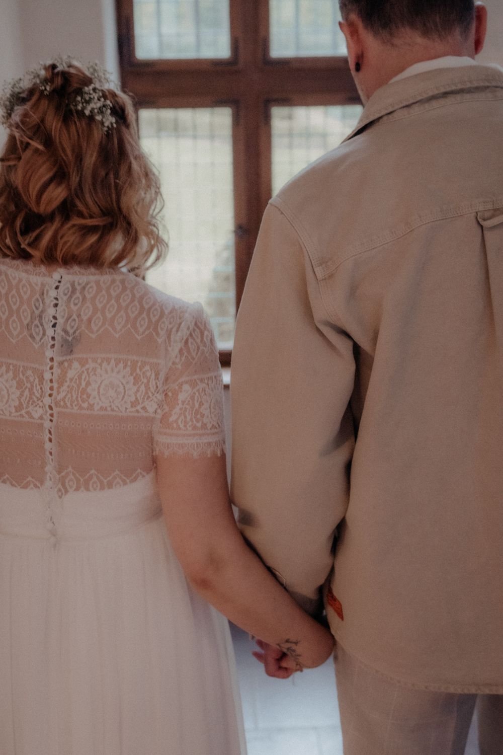 Ein Paar hält sich an den Händen, beide tragen helle, elegante Kleidung, im Hintergrund ein Fenster mit Gitterstäben.