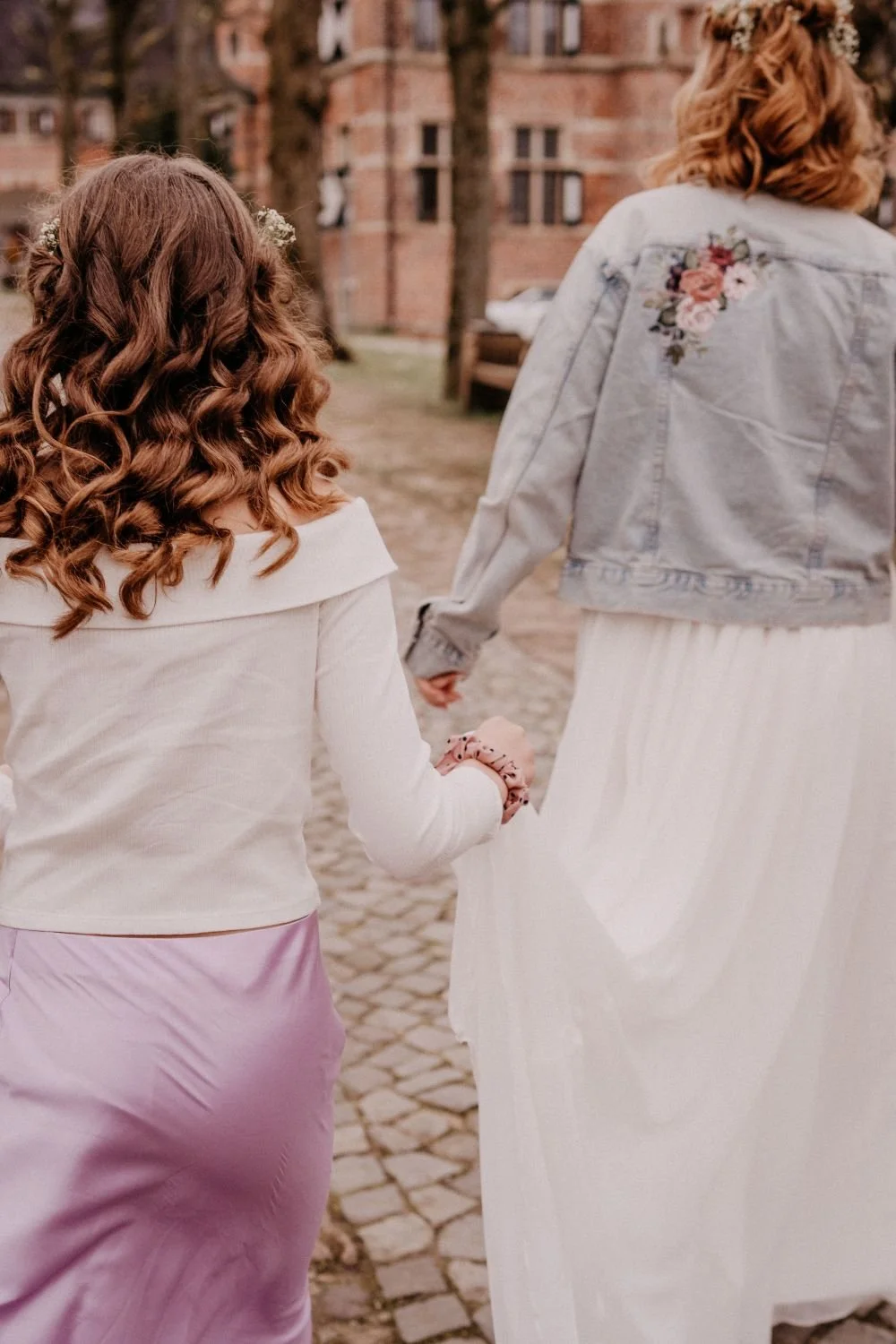 Zwei Frauen halten Händchen und gehen auf einer gepflasterten Straße, eine trägt ein weißes Kleid mit Blumenmuster auf der Rückseite, die andere trägt eine weiße Jacke mit einem Blumenmuster auf der Schulter.