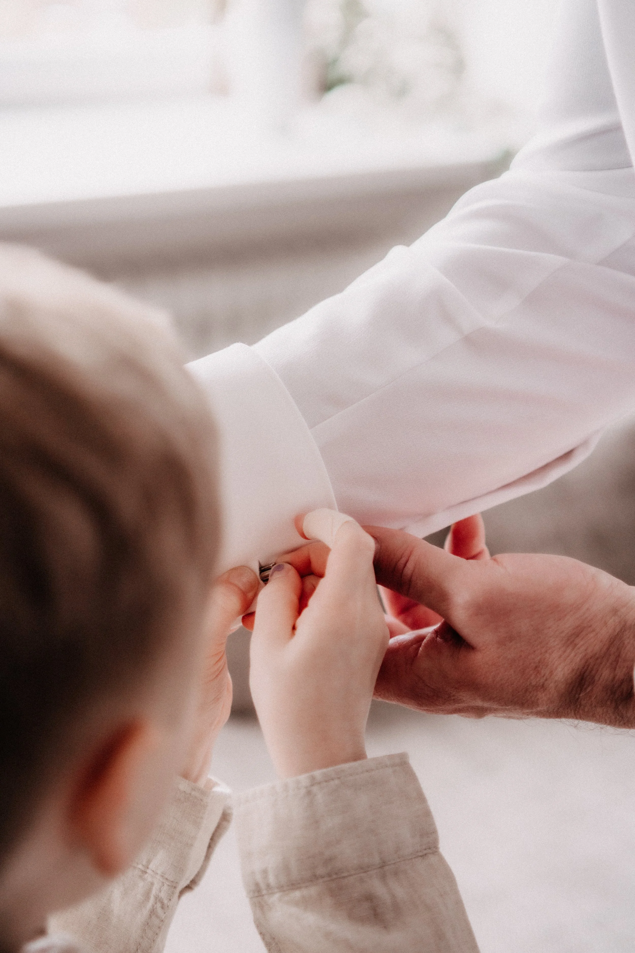 Nahaufnahme von einer Person, die einem Kind hilft, eine weiße Jacke zuzuknöpfen, im Innenraum mit natürlichem Licht.