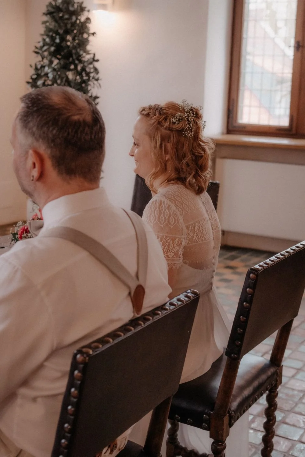 Braut und Bräutigam sitzen während einer Zeremonie in einem Raum mit Holzfenster, Holzboden und einem kleinen Weihnachtsbaum im Hintergrund.
