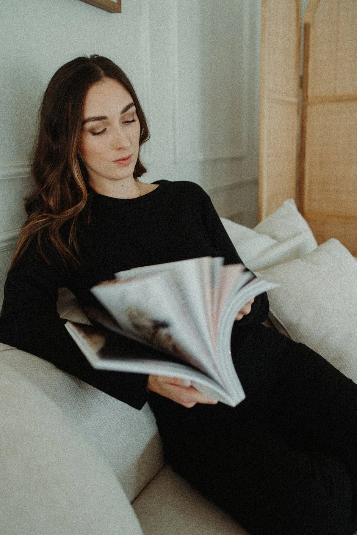 A young woman with brown hair reading a magazine on a beige couch in a cozy room.