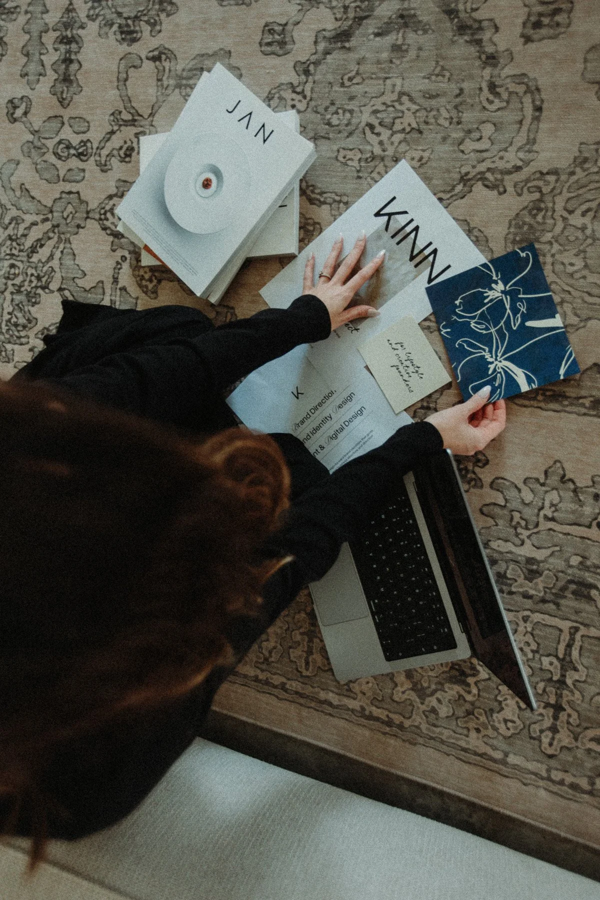 Top-down view of a person sitting at a wooden table, surrounded by papers, books, gift bag, and a laptop, working on a creative project.