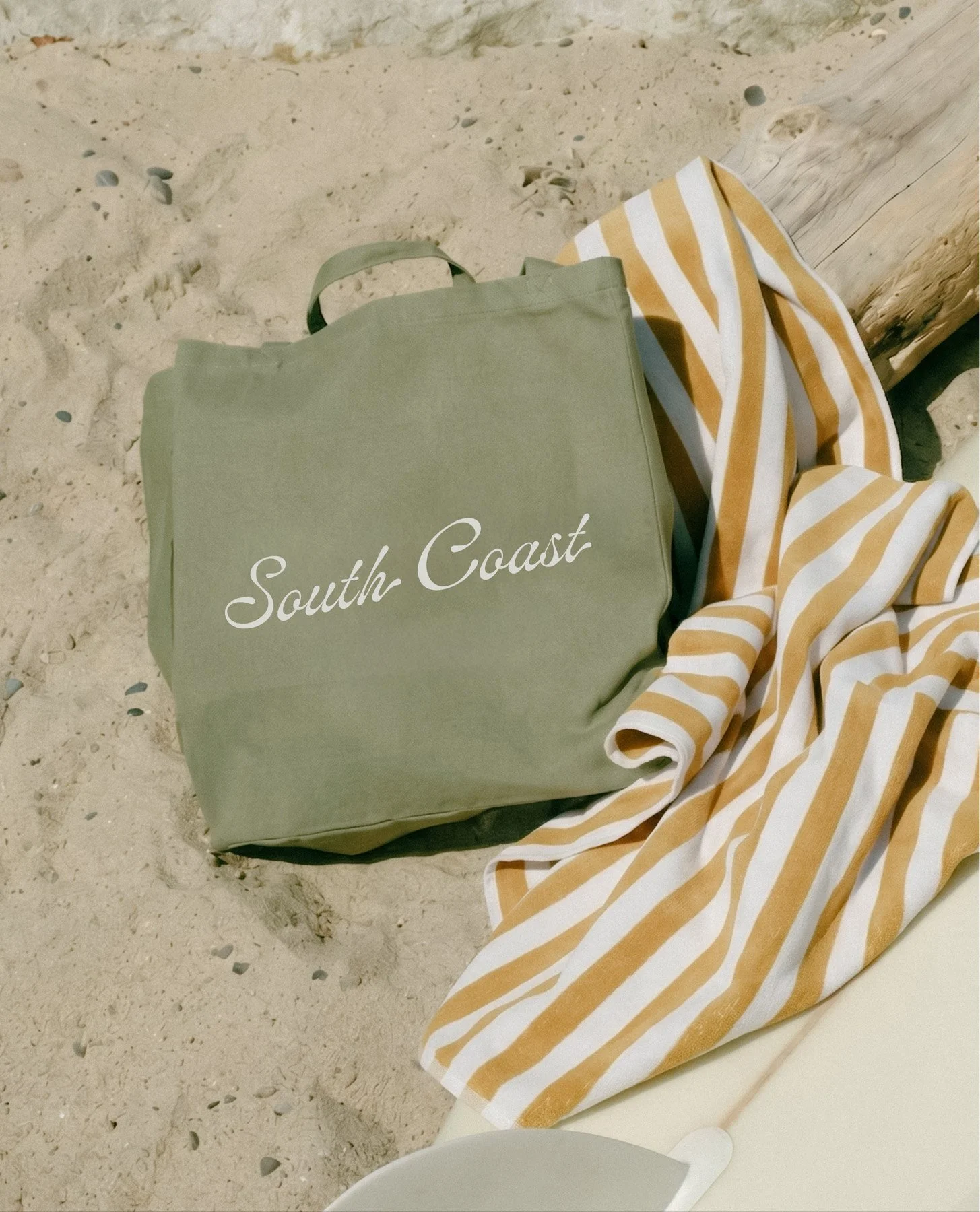 A green tote bag with the words 'South Coast' written on it. It is placed on the sandy ground next to a striped orange and white beach towel or blanket. Part of a wooden post or wall is visible in the background.