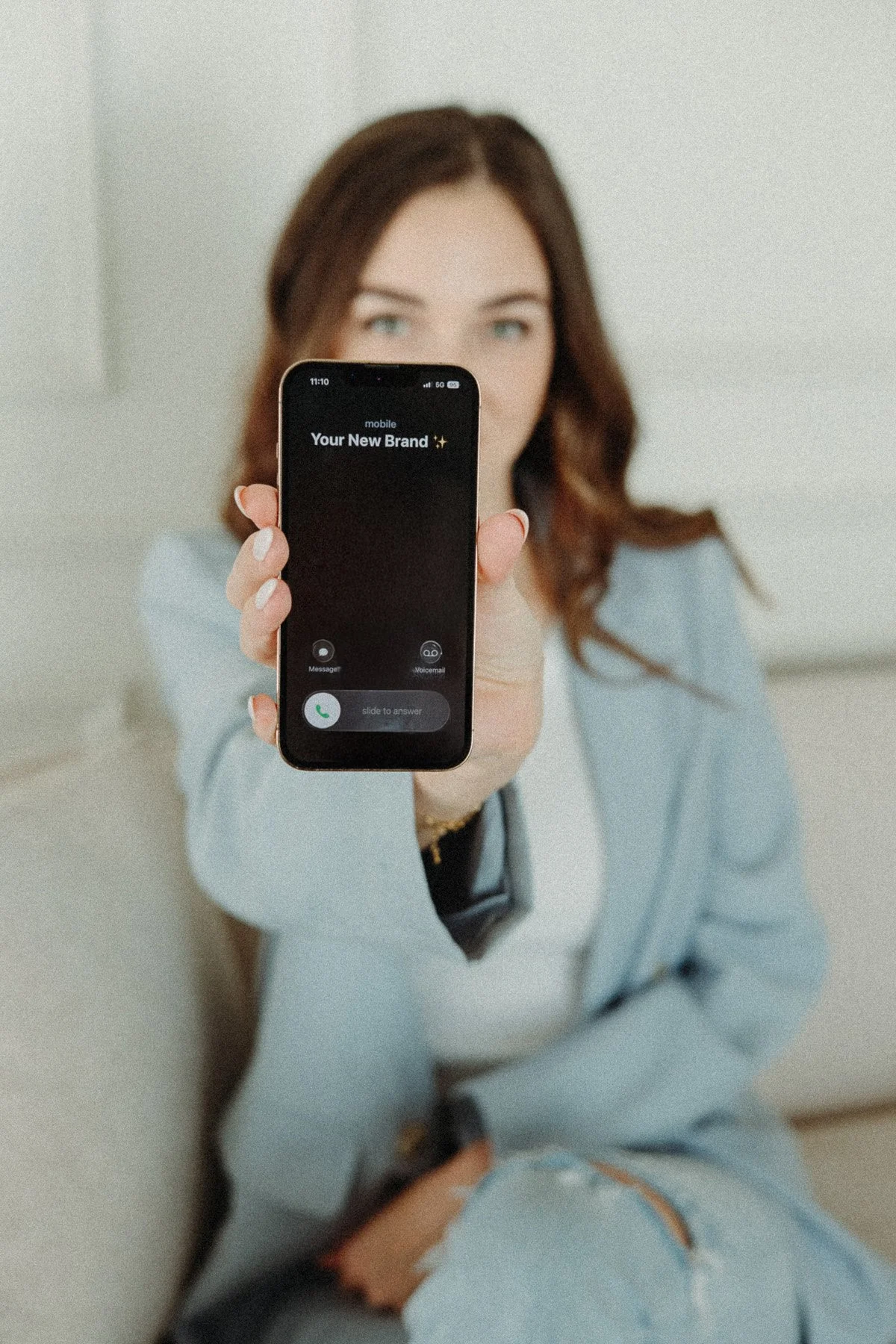 Woman with brown hair wearing a light blazer and ripped jeans, sitting on the floor, holding a smartphone towards the camera, displaying an incoming call with the message 'Your New Brand ✨'.