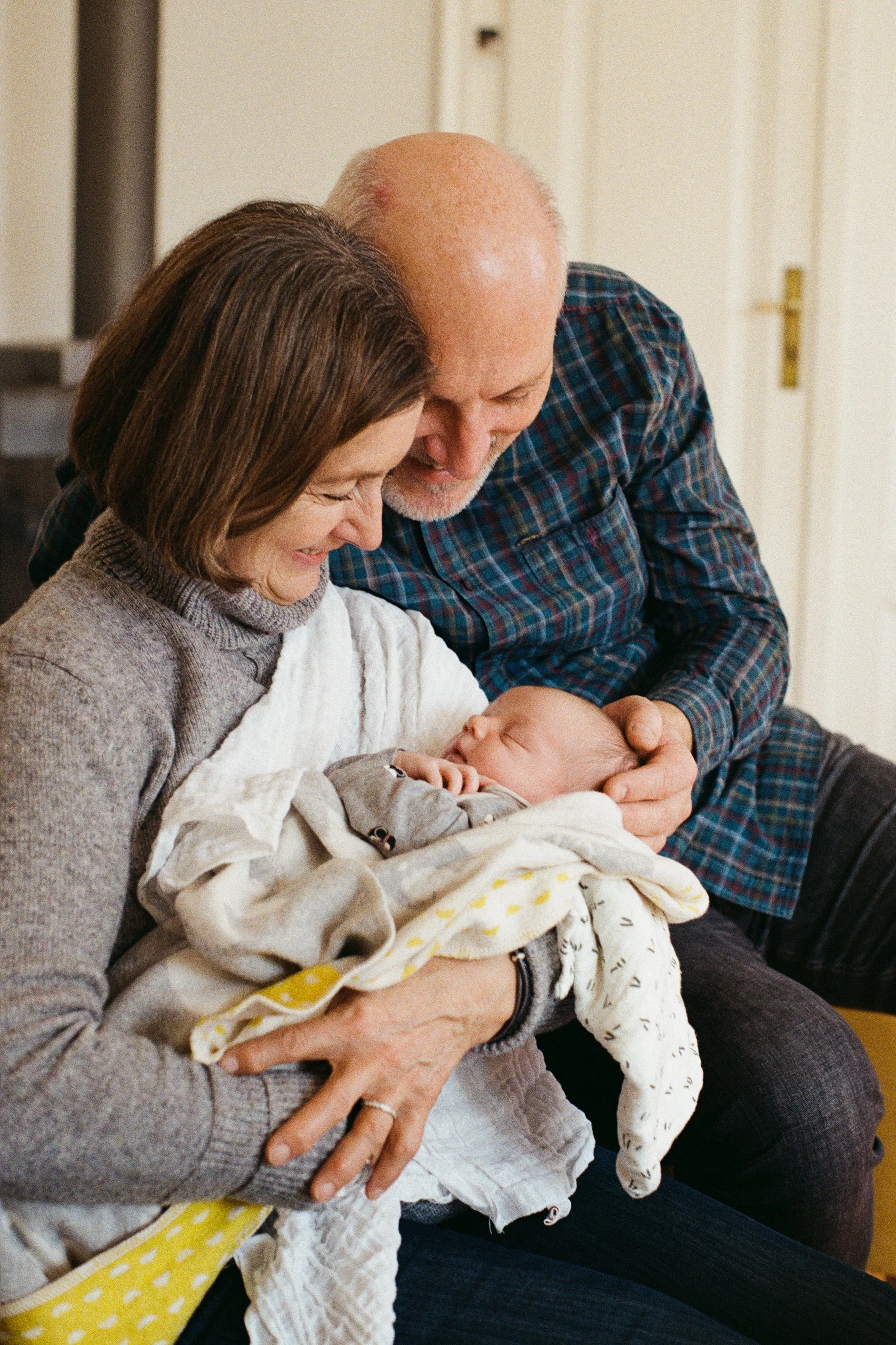 An older woman and man hold a sleeping baby, smiling and looking affectionately at the baby.