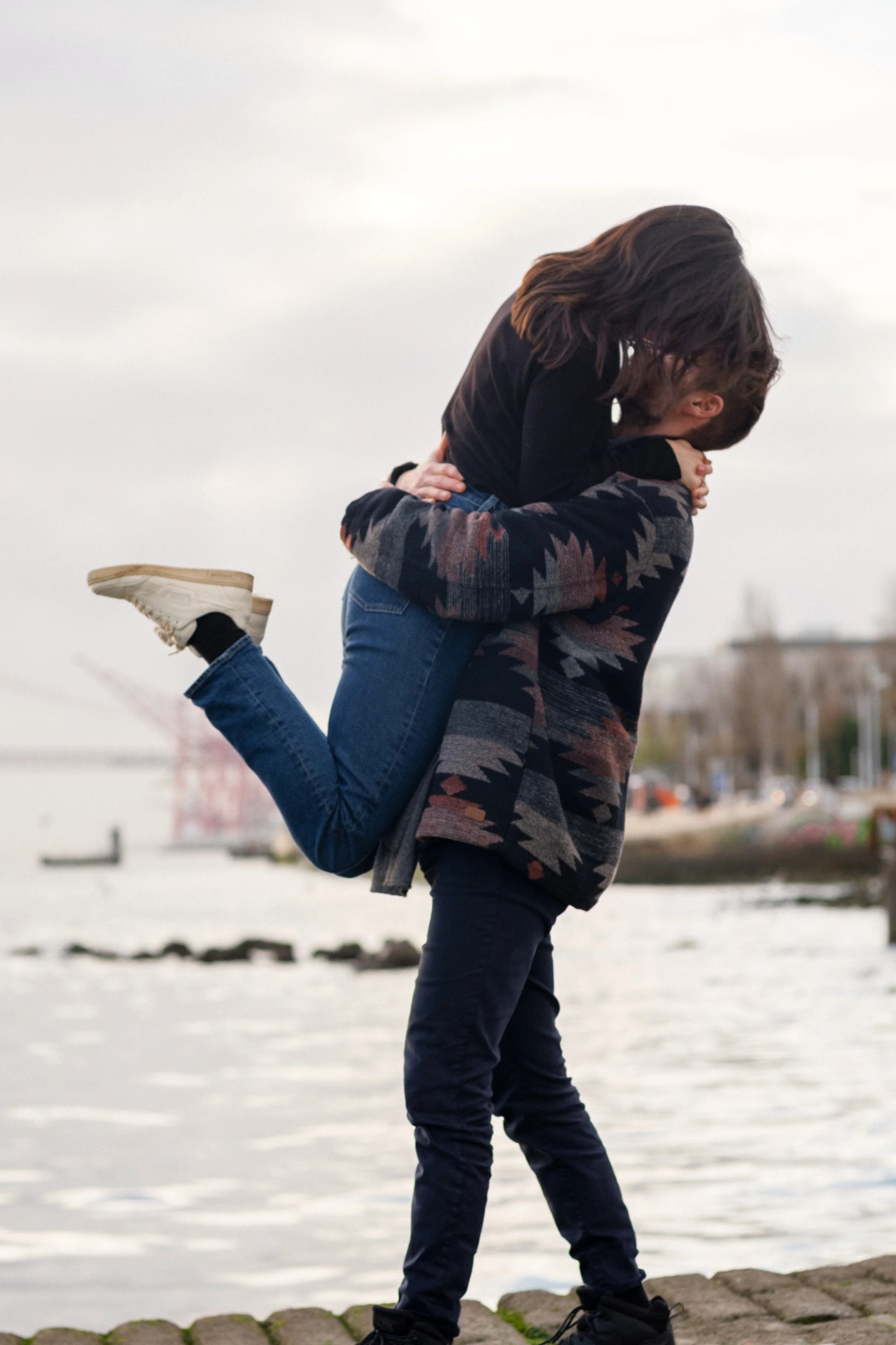 A couple by the water, with the man lifting the woman in his arms, kissing her, and smiling, during daytime with cloudy skies.
