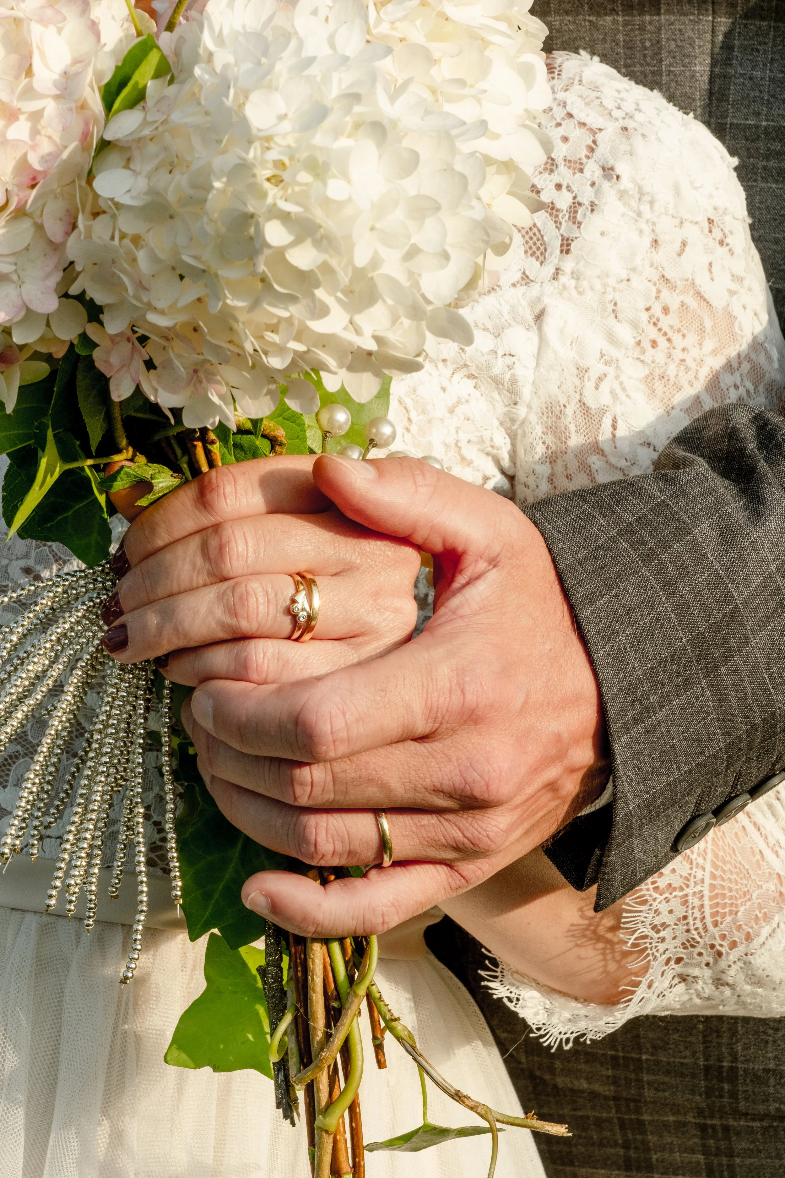 Close-up of a bride and groom holding a bouquet of white and pale pink flowers and greenery. The bride is wearing a lace dress and the groom a gray suit with a dark shirt. Wedding rings are visible on their hands.