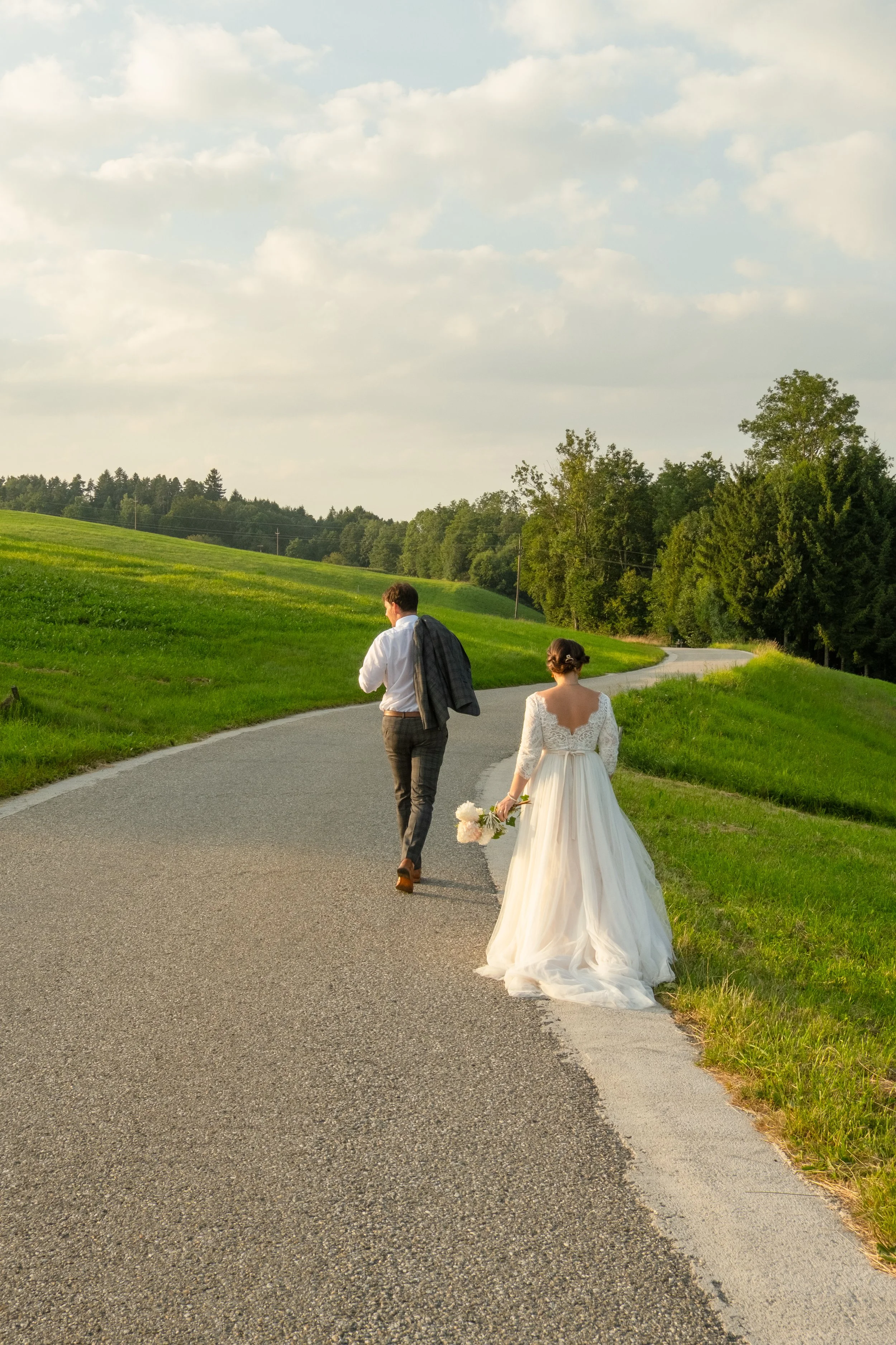 A bride and groom walking along a winding country road, the bride holding a bouquet of flowers, surrounded by green fields and trees.
