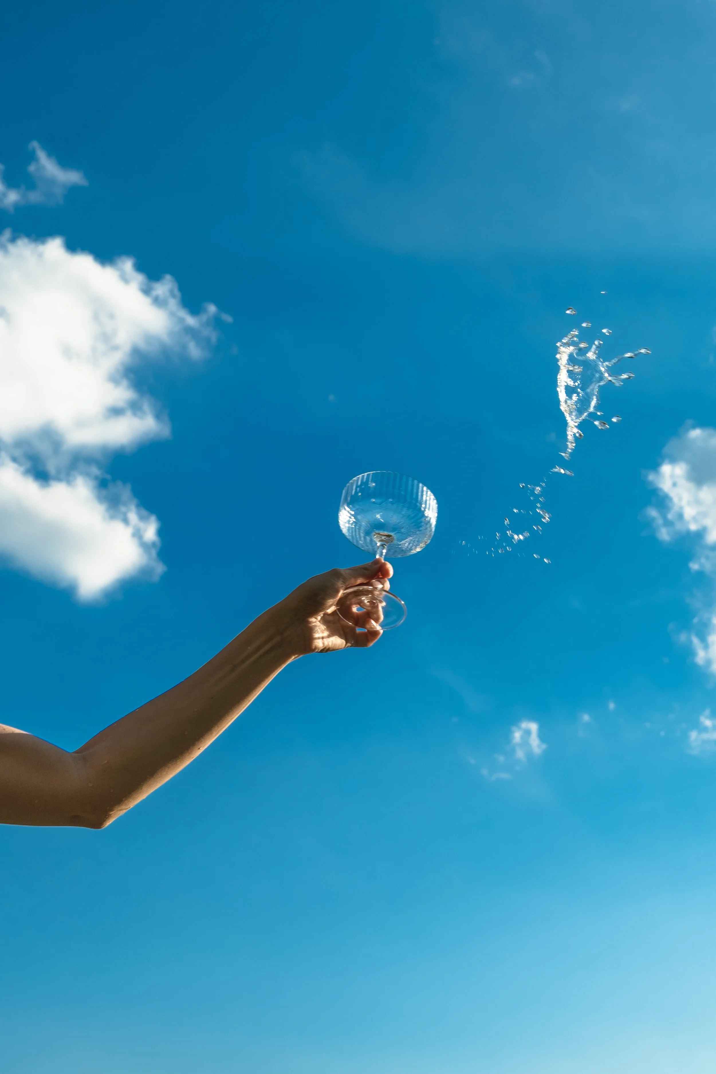A person holding a glass with water against a blue sky with a few clouds, water splashing out of the glass.
