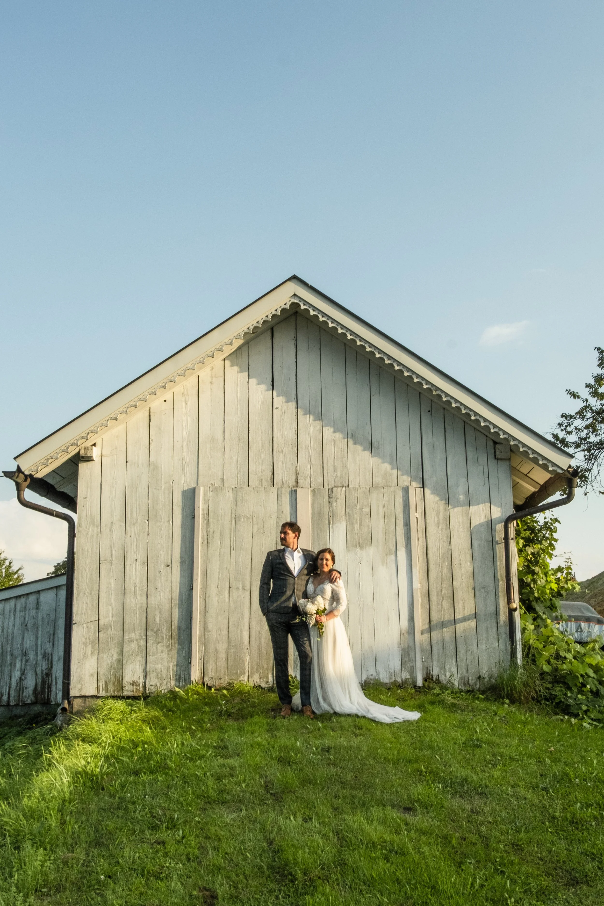 A bride and groom standing together outside a rustic wooden barn, the groom in a checkered suit and the bride in a long white wedding dress, holding a bouquet, during sunset.