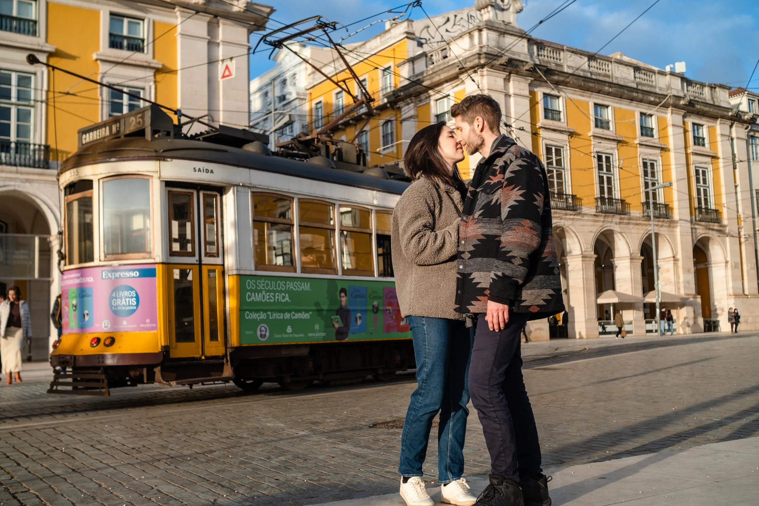 A couple standing close together, about to kiss, in front of a vintage yellow tram on a cobblestone street in an urban area with yellow and white historic buildings.