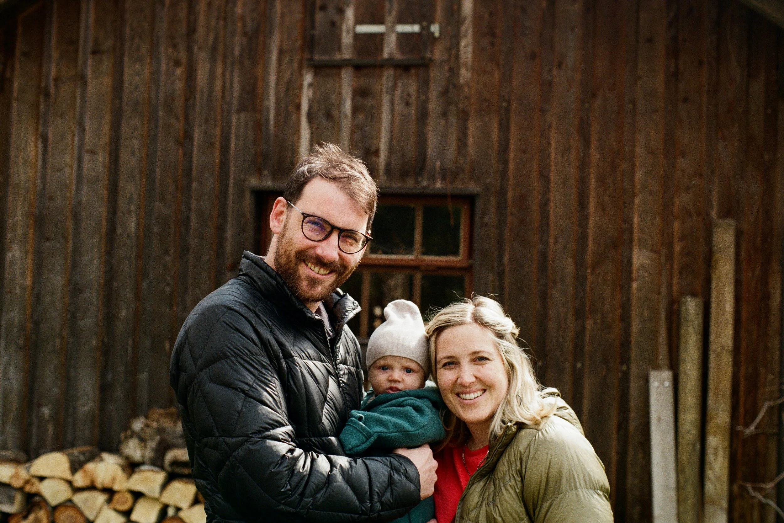 A smiling young family of three, with a man, woman, and baby, standing outdoors in front of a wooden shed with wood logs stored outside, during daytime.