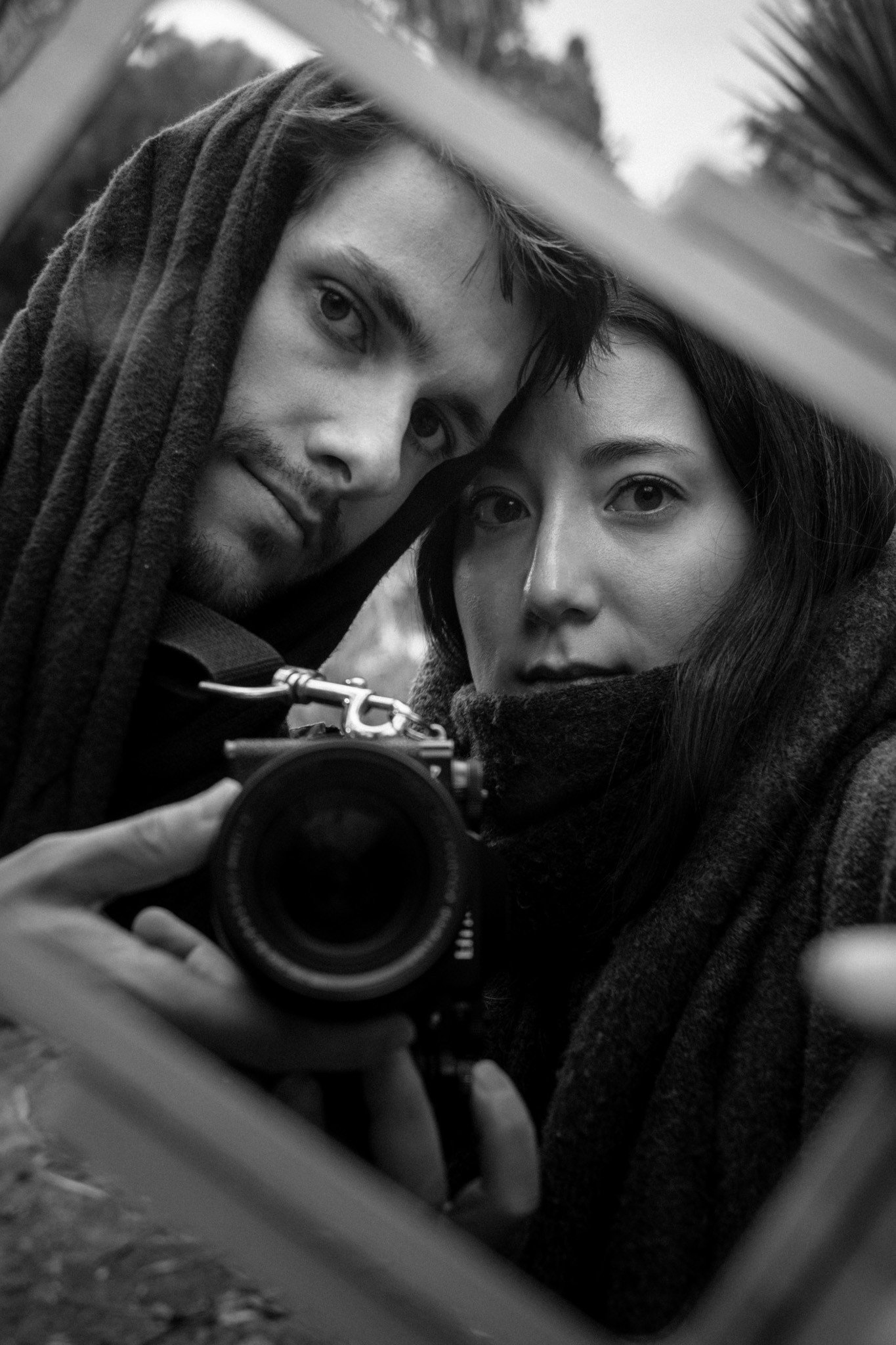 Two people taking a selfie outdoors, one man with a beard and dreadlocks, and a woman with long dark hair, both looking into the camera with serious expressions, using a camera as the mirror for the selfie.