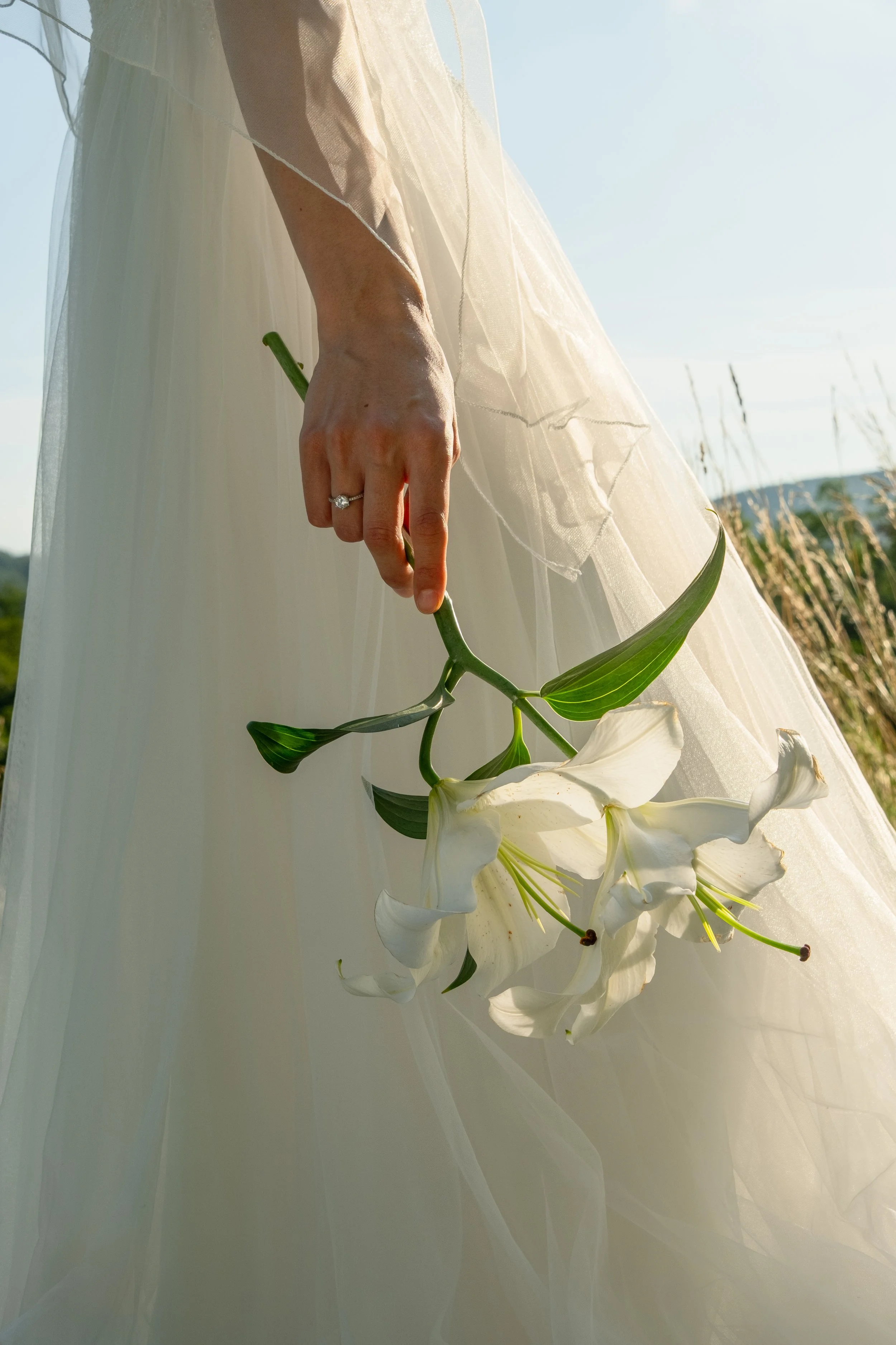 A person in a white dress holding a bouquet of white lilies, with part of their hand and dress visible in a natural outdoor setting.