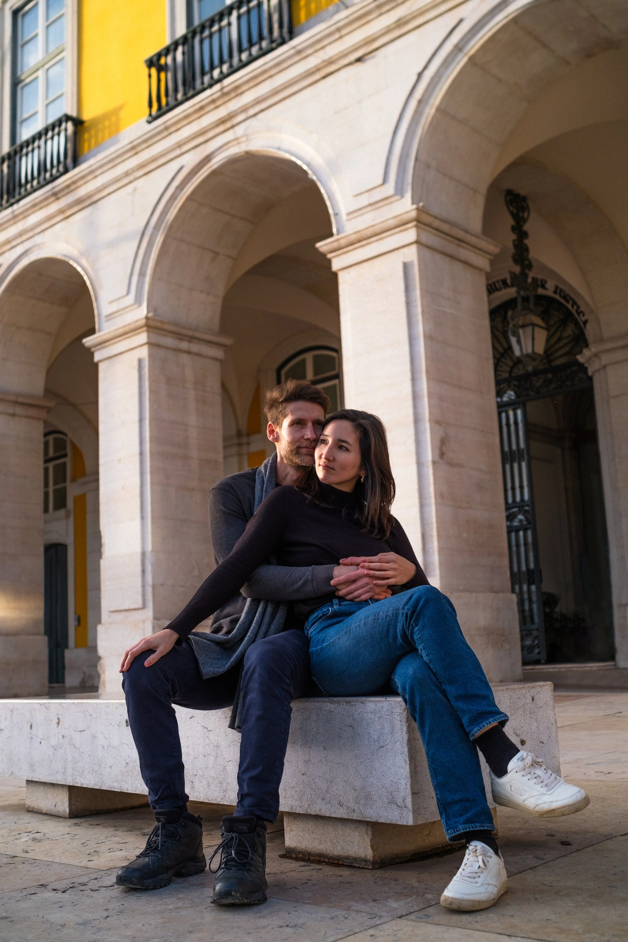 A young couple sits on a stone bench in an outdoor courtyard with classical architecture, including large arches and decorative ironwork. The man has short hair and a beard, wearing a gray jacket and dark pants, while the woman has shoulder-length dark hair, wearing a black top and blue jeans. They appear to be spending time together, with a warm, intimate atmosphere.