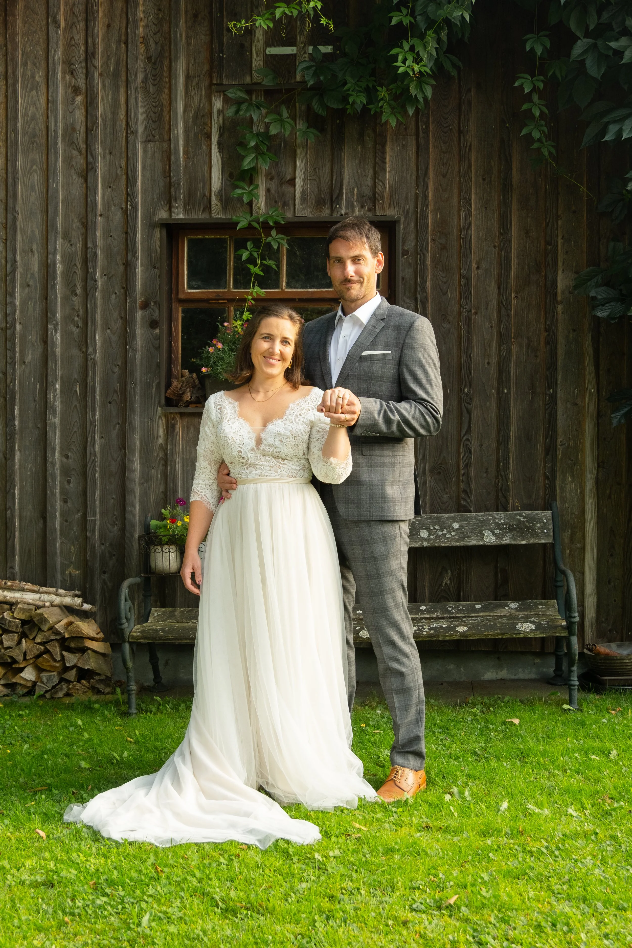 A newlywed couple holding hands outdoors in front of a rustic wooden wall with a window. The woman wears a white lace wedding dress with a flowing skirt, and the man wears a gray plaid suit. There are green plants and a bench behind them.