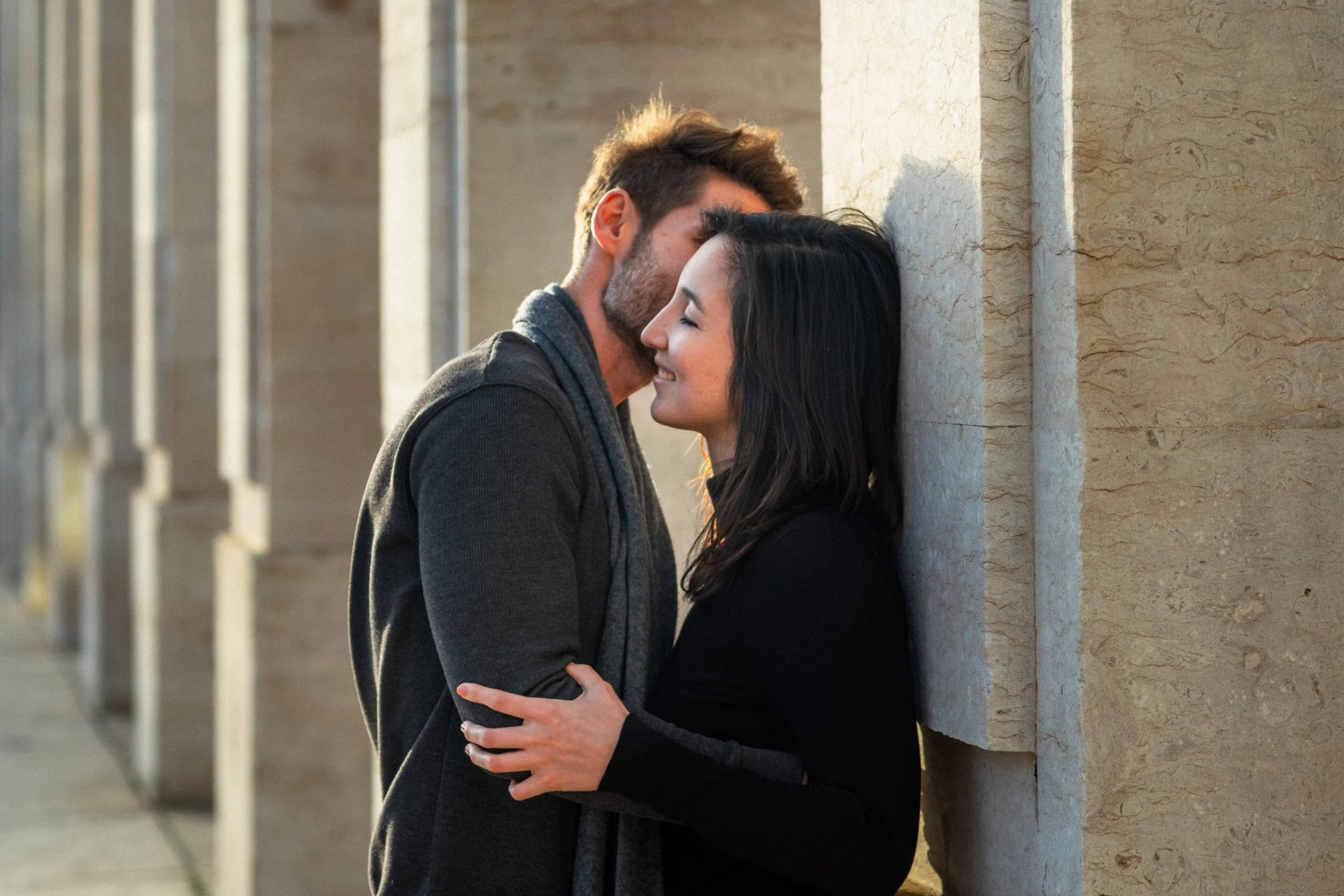 A couple sharing an intimate moment, with the man whispering into the woman's ear as she smiles with eyes closed, leaning against a wall.
