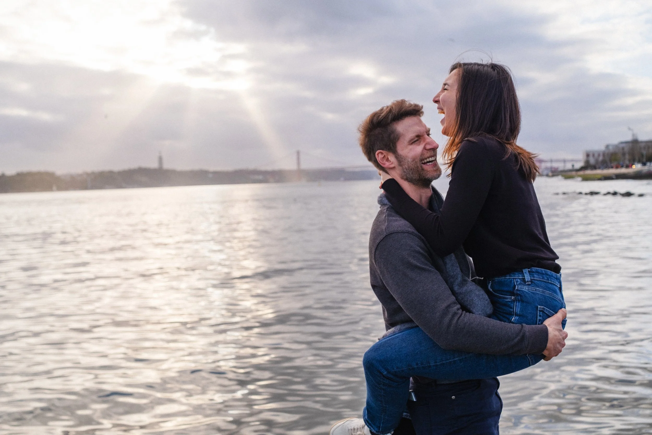 A man lifts a woman by the water with the San Francisco Bay Bridge in the background, both smiling and looking at each other.
