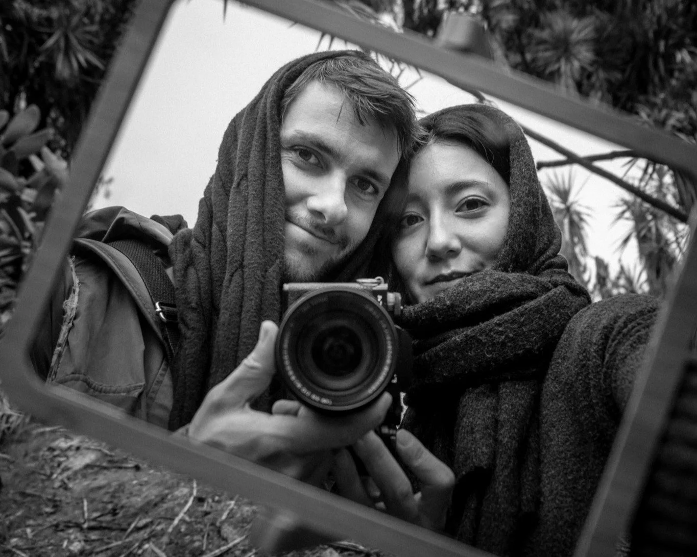 Black and white photo of a man and woman taking a selfie together outdoors, using a mirror to capture the shot, both wearing hooded scarves, trees in the background.