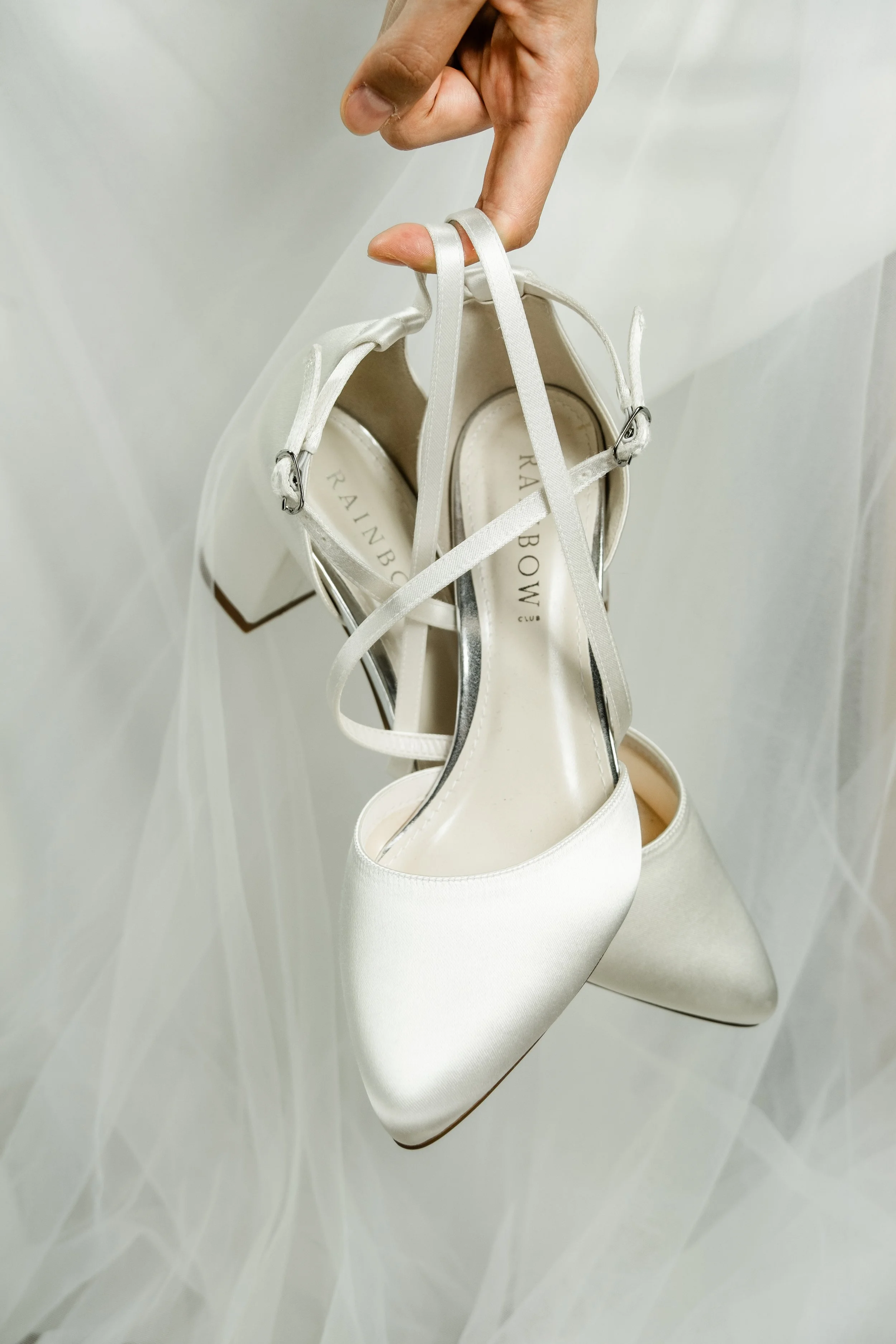 A hand holding a pair of white satin high-heeled shoes with pointed toes, hanging against a white background.