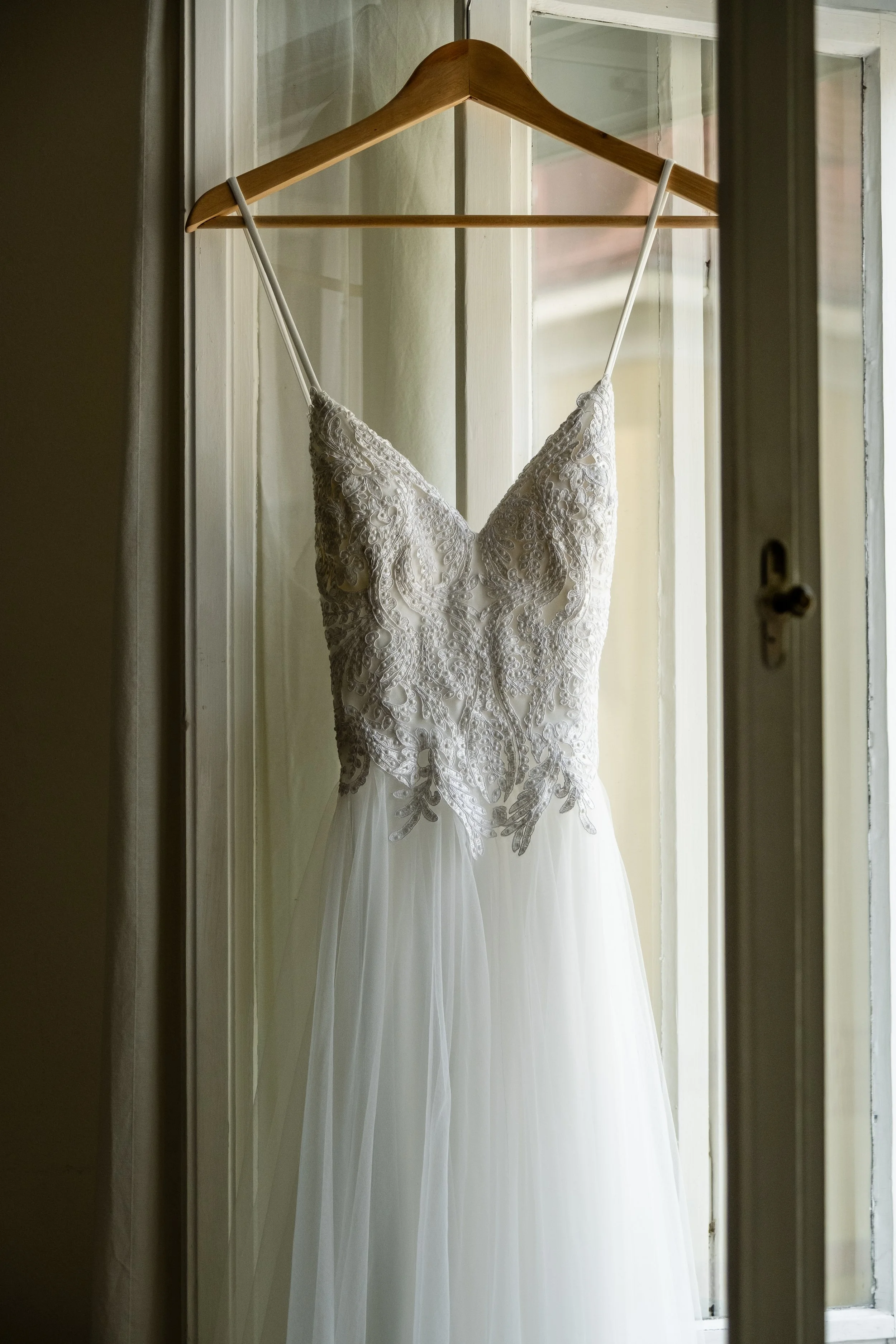 Elegant white wedding dress with lace bodice hanging on a wooden hanger in front of a window.