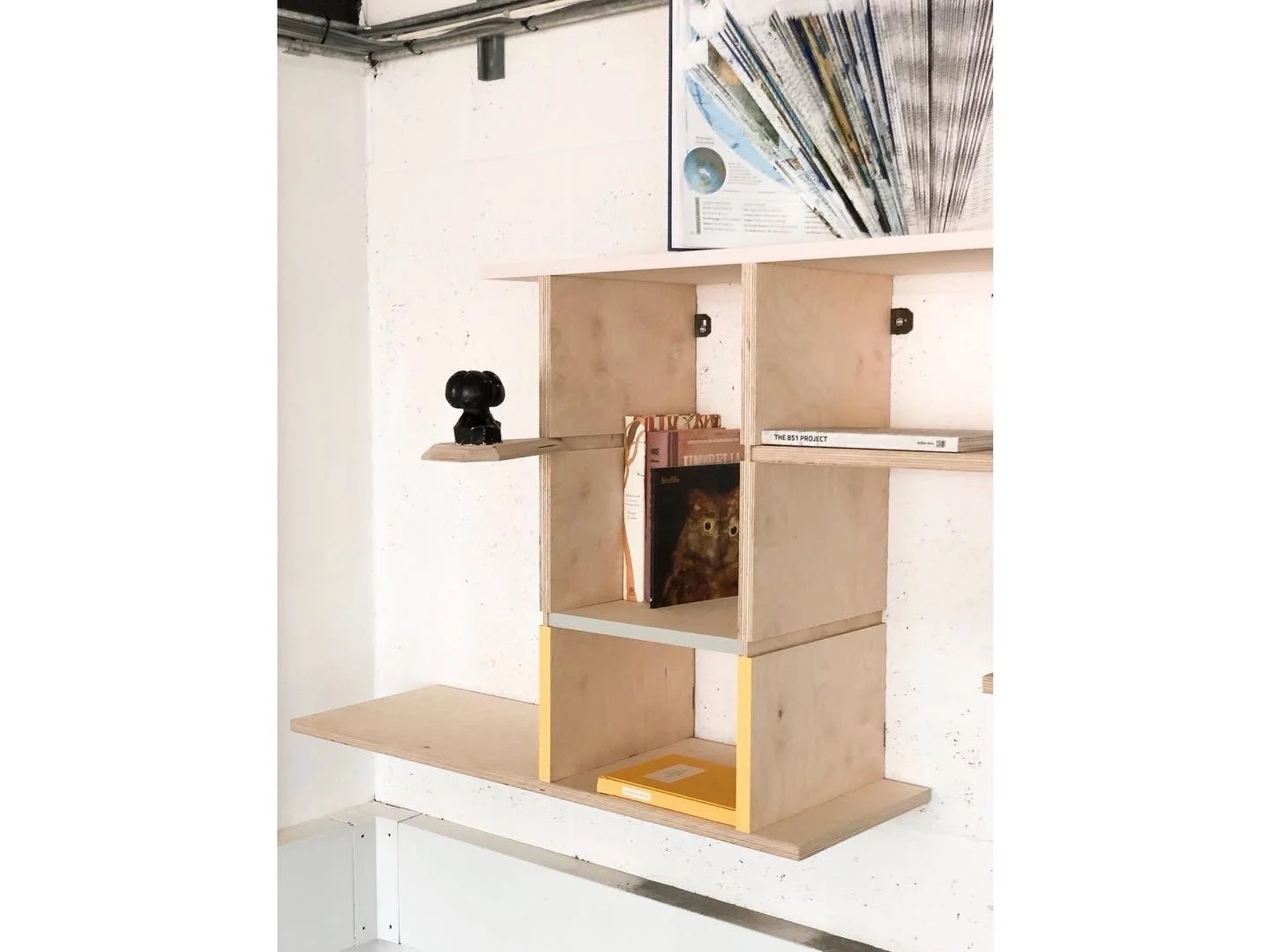 Wooden bookshelf with multiple compartments, holding books, a black decorative object, and a yellow folder, mounted on a white wall.