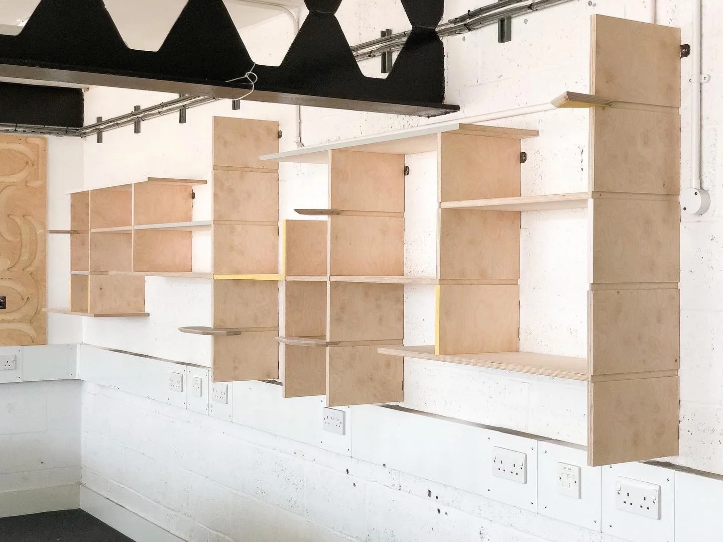 Wooden modular shelves attached to a white brick wall, with black fabric and metal fixtures on the ceiling, visible electrical outlets along the wall.