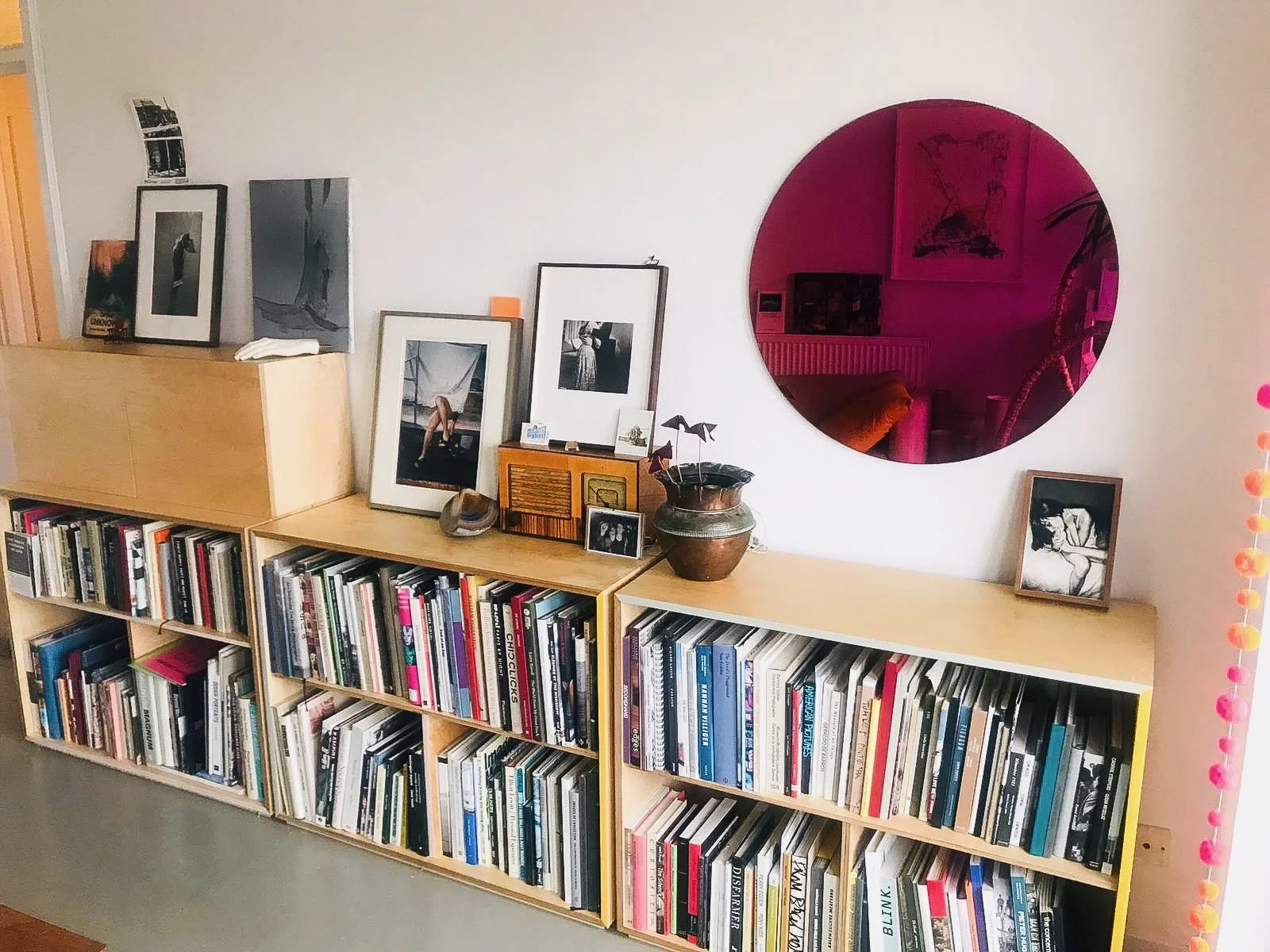 A wooden bookshelf filled with books and framed photographs, topped with artwork, a small plant, and decorative items, situated in a room with a large circular mirror reflecting part of a cozy living space.