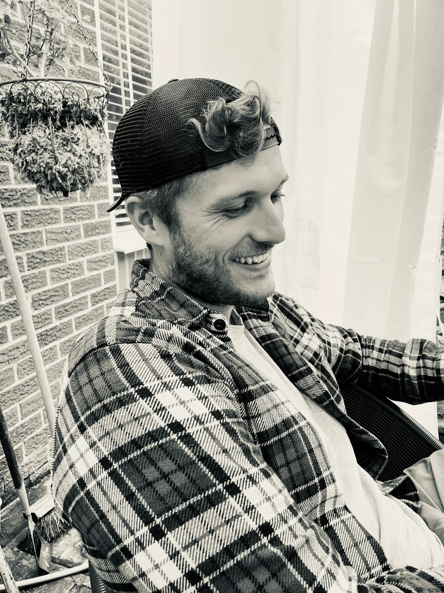 A young man with a beard, wearing a baseball cap backwards, plaid shirt, and light sweater, smiling while sitting outdoors near a brick wall and window with blinds.