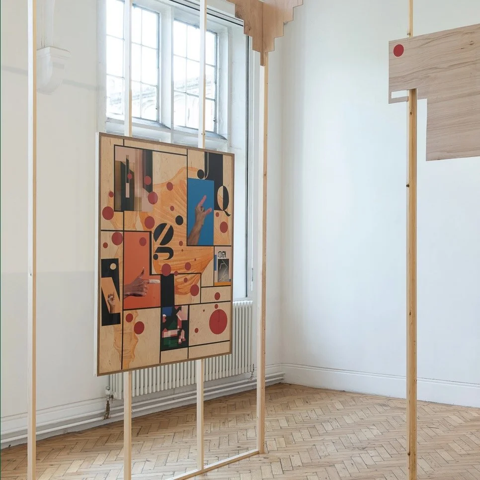 A colorful abstract art piece with illustrations of hands, geometric shapes, and circles, displayed in a minimal gallery with wooden framing and a large window in the background.