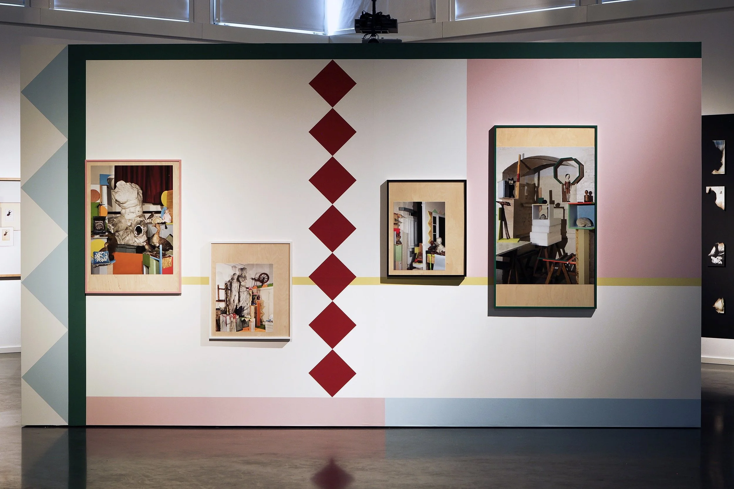 Art exhibit featuring four framed photographs of installations or sculptures on a colorful geometric wall with red diamonds and pastel patterns.