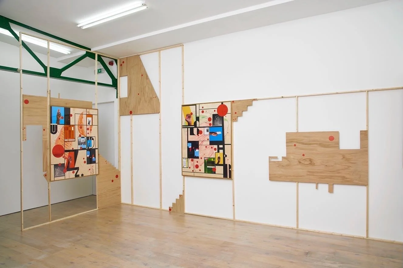 An art installation on a gallery wall featuring geometric artwork with wood, black lines, and colorful red, orange, blue, yellow, and green accents. The wall includes sections of wood panels and open framing.