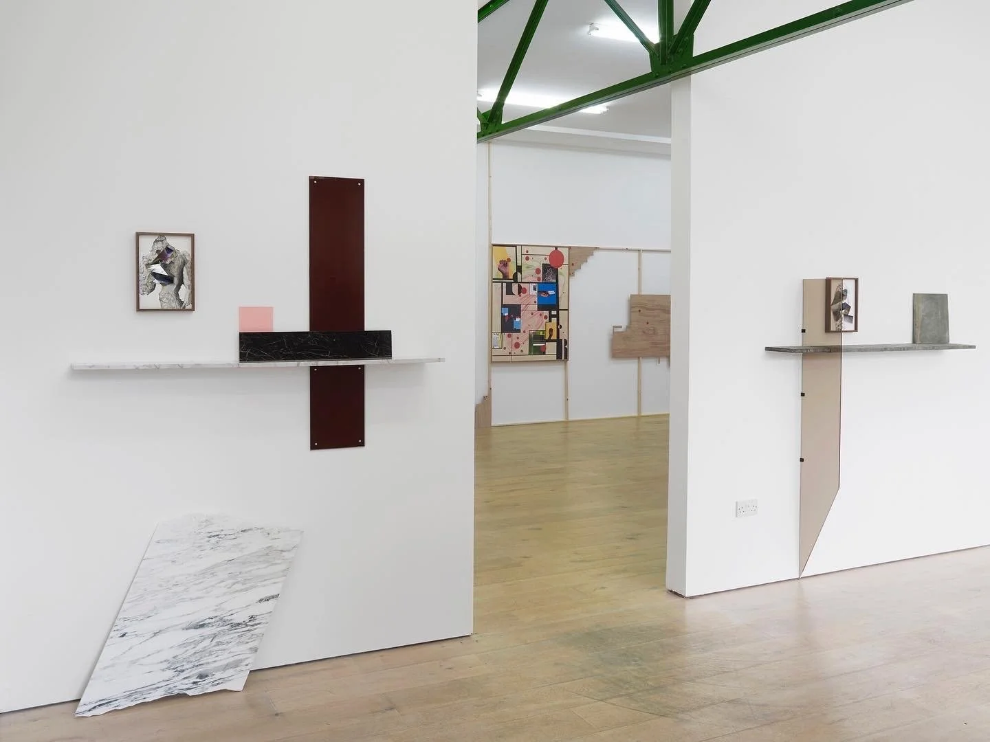 Minimalist art display with small framed artwork and textured materials arranged on white walls in an art gallery with light wood flooring and industrial ceiling structure.