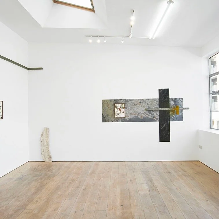 Empty art gallery room with white walls, wooden floor, and modern artwork on the wall. There is a sculpture in the corner, a large rectangular marble piece on the wall, and contain lights on the ceiling. A window is visible on the right side.