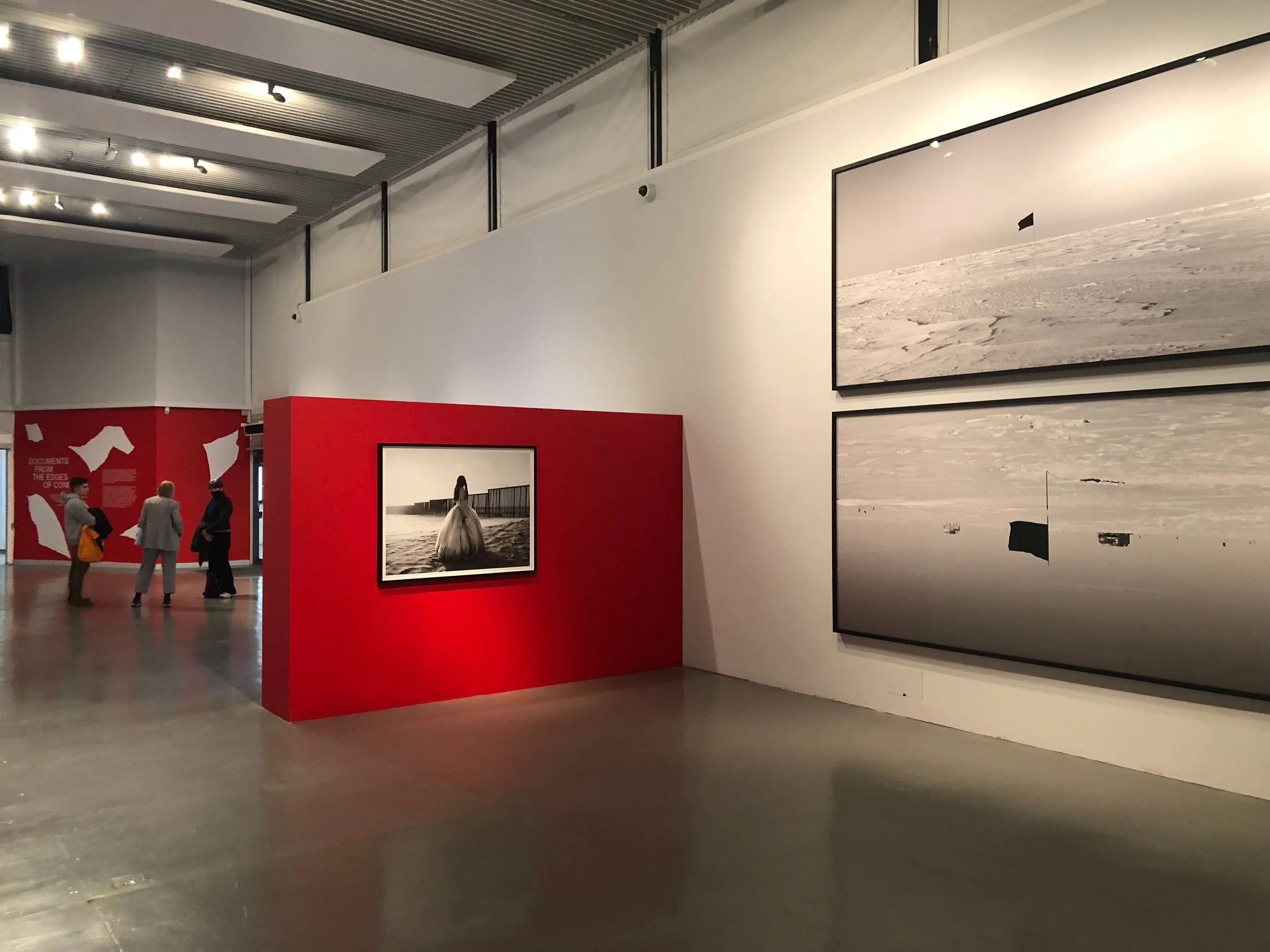An art gallery with black and white photographs on plain white walls, a red partition with a framed photo of a woman in a dress by the water, and a red wall with white text and illustrations in the background. Three people are talking in the distance