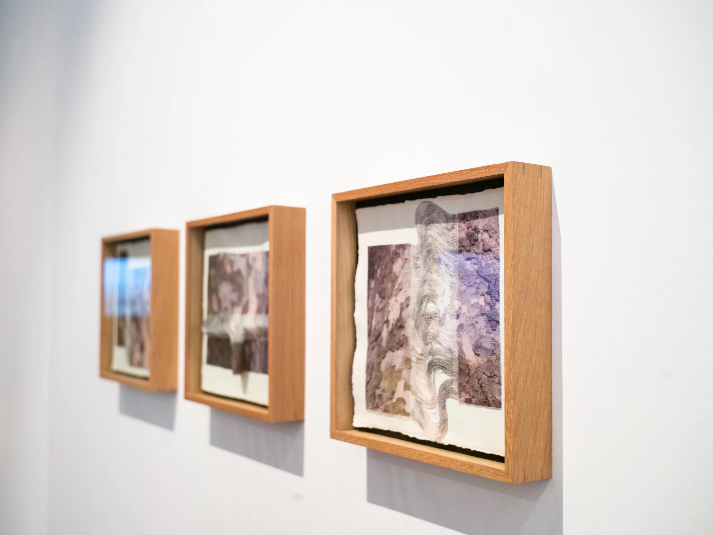 Three framed abstract artworks with textured paper in wooden frames hanging on a white wall.