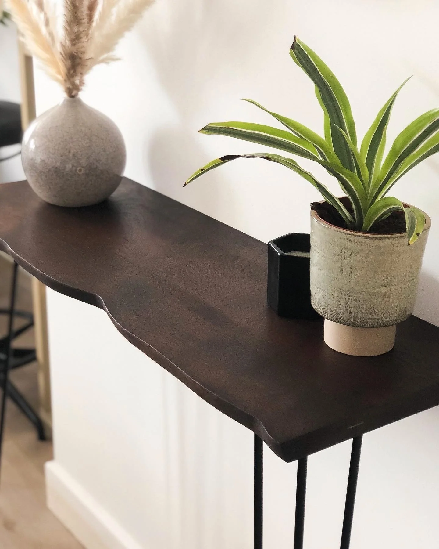Decorative wooden ledge with a round textured vase holding pampas grass and a potted plant, along with a small black container, against a white wall.