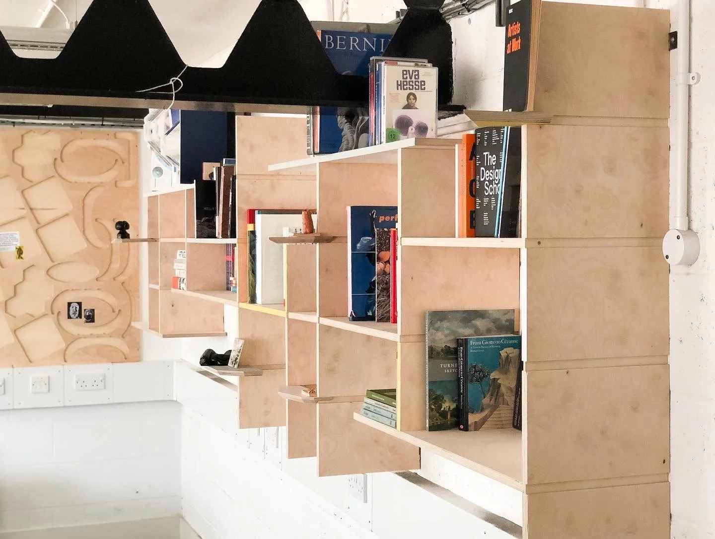 A wooden bookshelf with multiple compartments displaying books in an indoor setting. The bookshelf is partially filled with books, with some placed vertically and others horizontally. The background includes a wall with electrical outlets and a decor