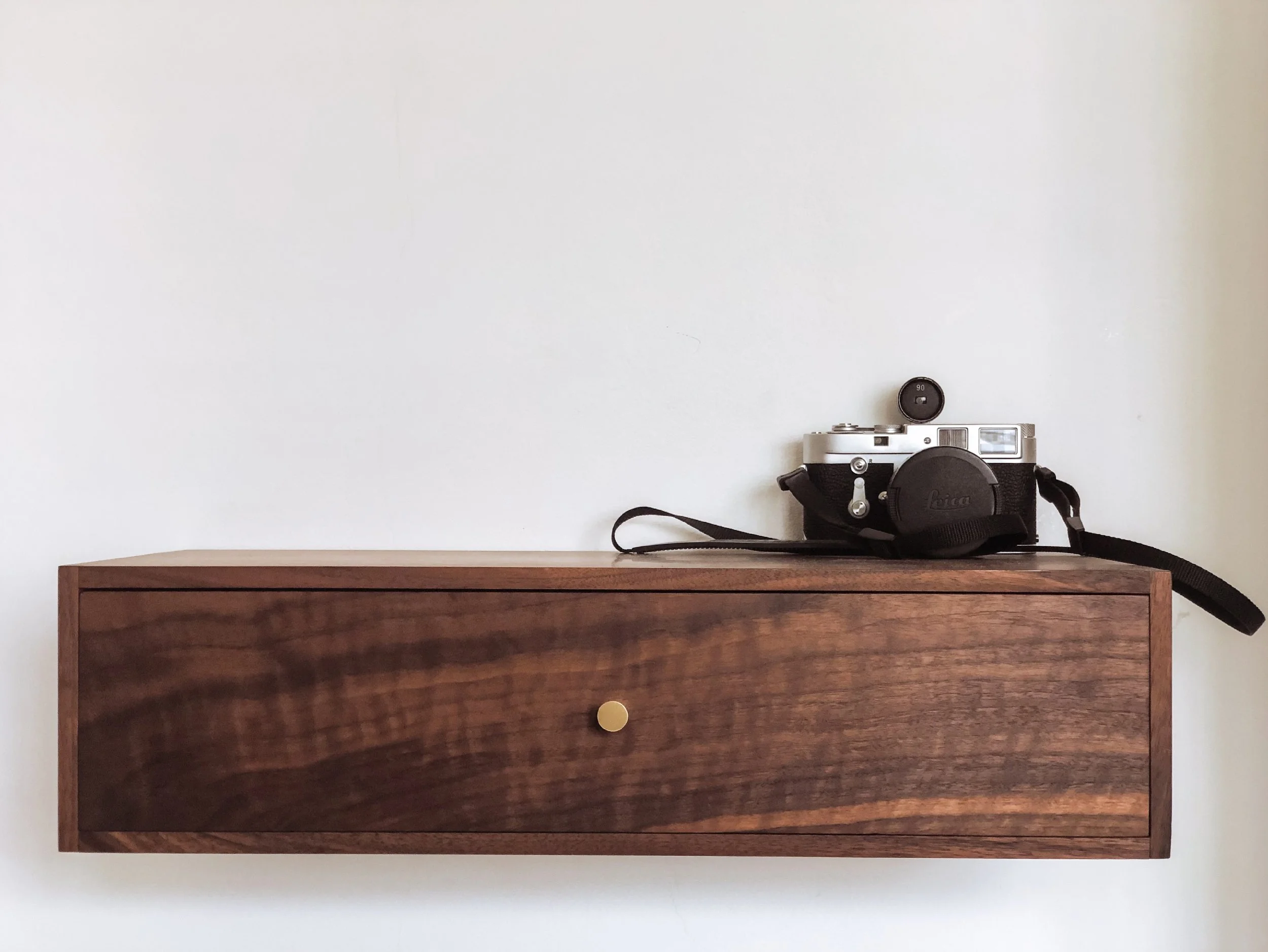 A vintage Leica camera with a lens cap on, resting on a wooden shelf against a plain white wall.