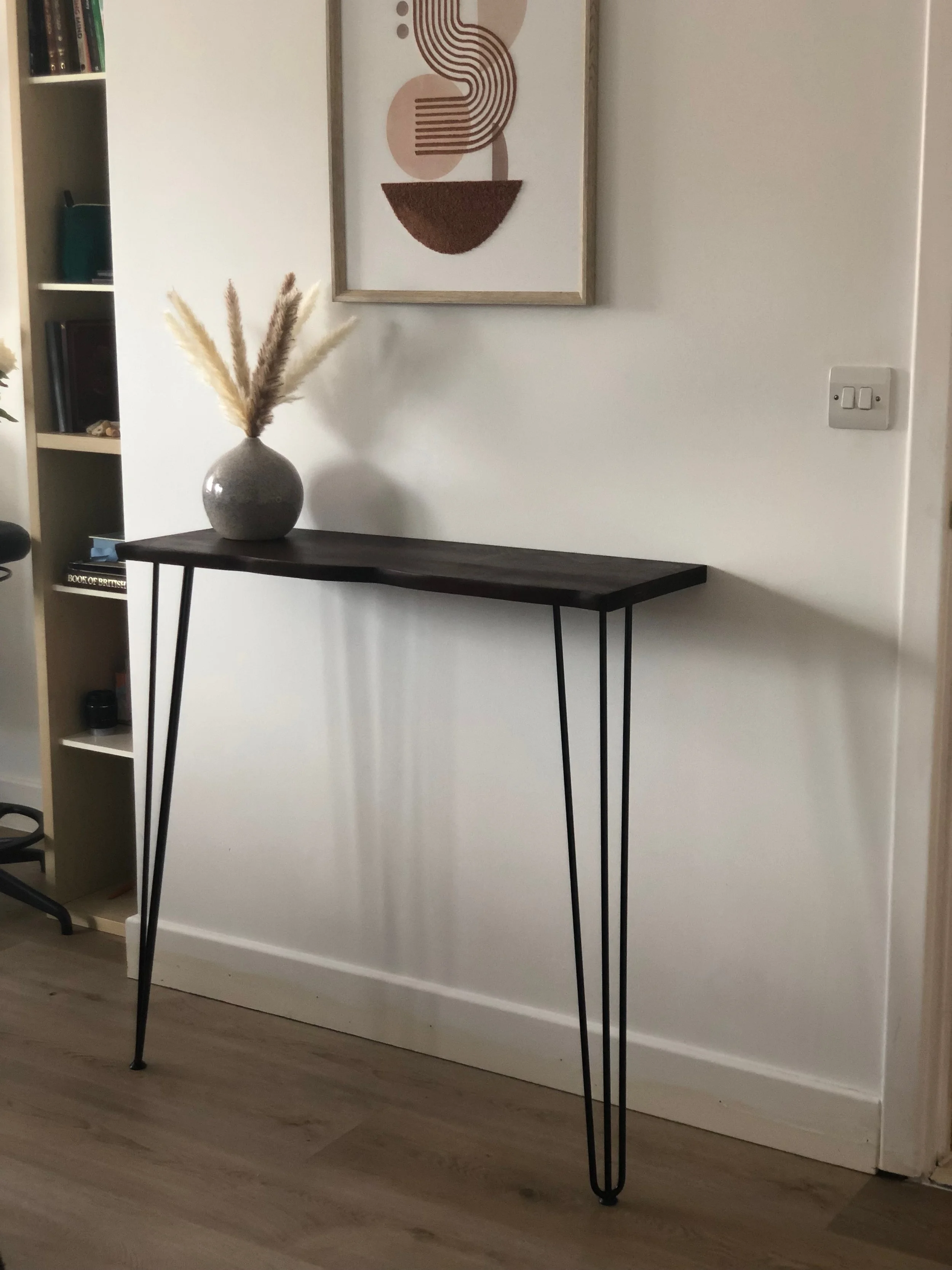 A dark wooden console table with metal hairpin legs against a white wall, decorated with a gray vase holding pampas grass. Above the table is a framed abstract art print.