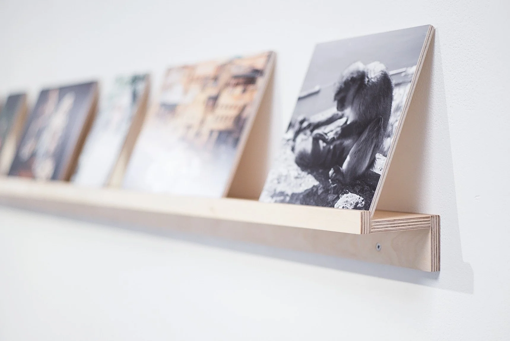 Black and white photograph of a woman sitting on rocks near the beach with her head bowed and her arms wrapped around her legs, displayed on a wooden shelf along with other blurred photographs.