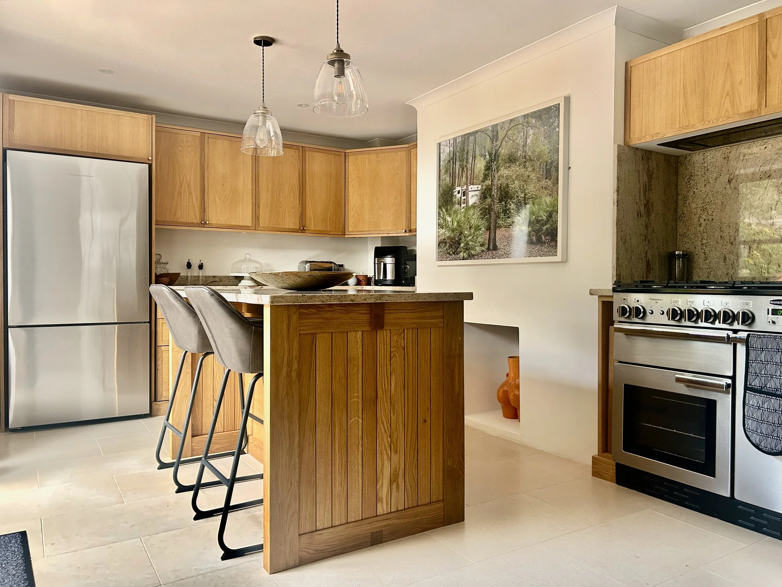 Modern kitchen with wooden cabinets, stainless steel refrigerator, and stove. Kitchen island with bar stools, framed nature photograph on wall, and decorative vases on a niche.