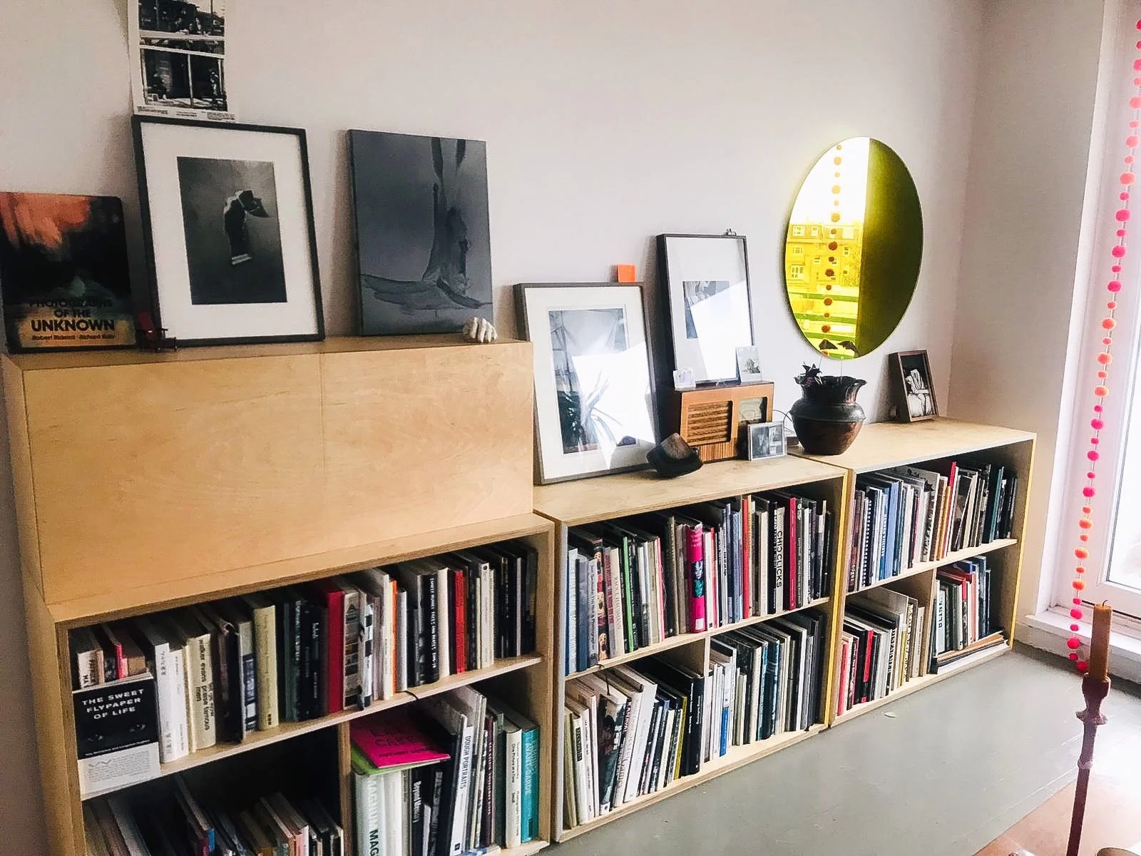 Wooden bookshelf against a white wall, topped with framed photographs and artwork, decorative objects, and a yellow oval mirror, with books on the shelves and a door with pink pom-pom garland to the right.