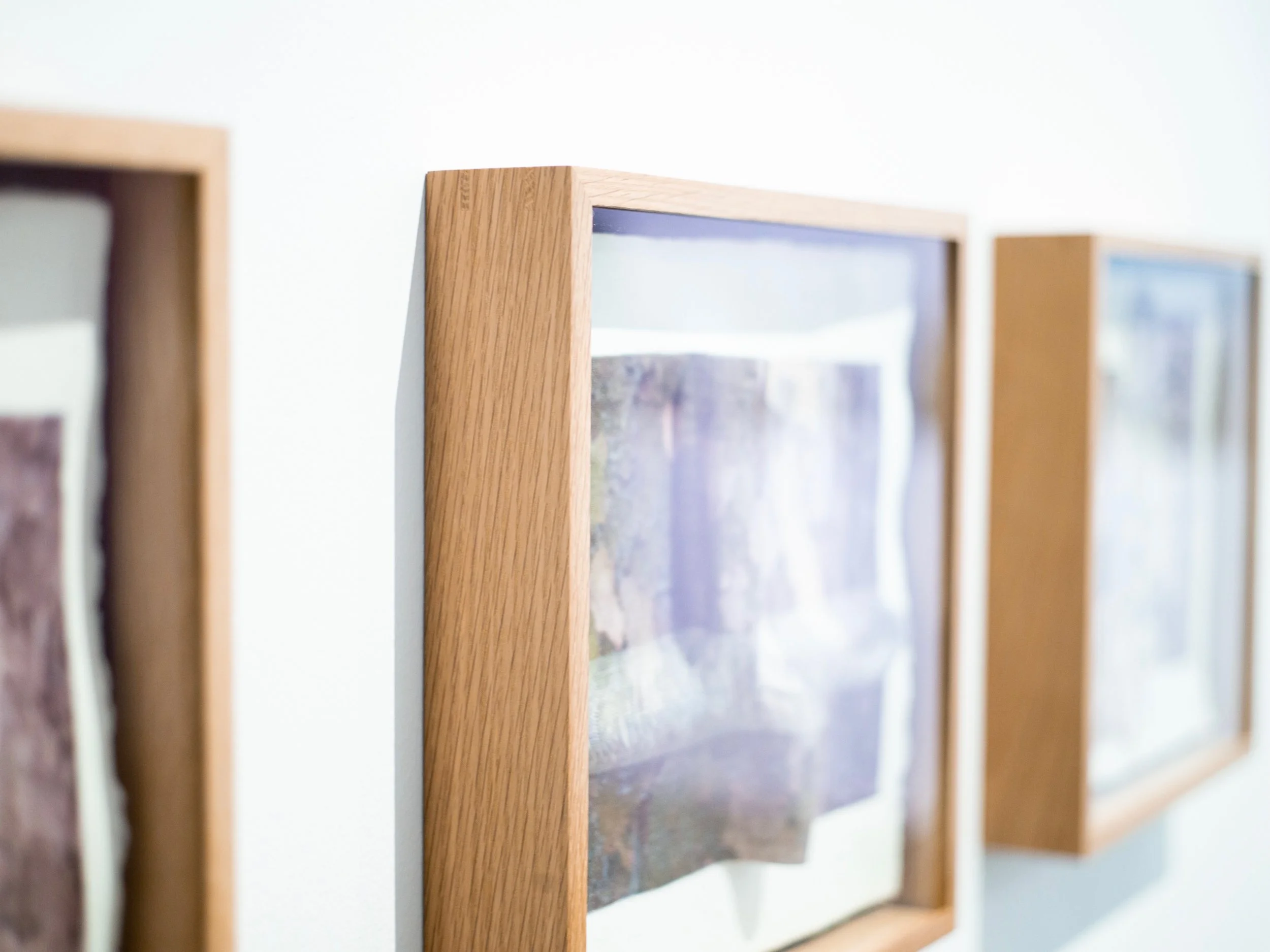Close-up of three framed abstract artworks with wooden borders hanging on a white gallery wall.
