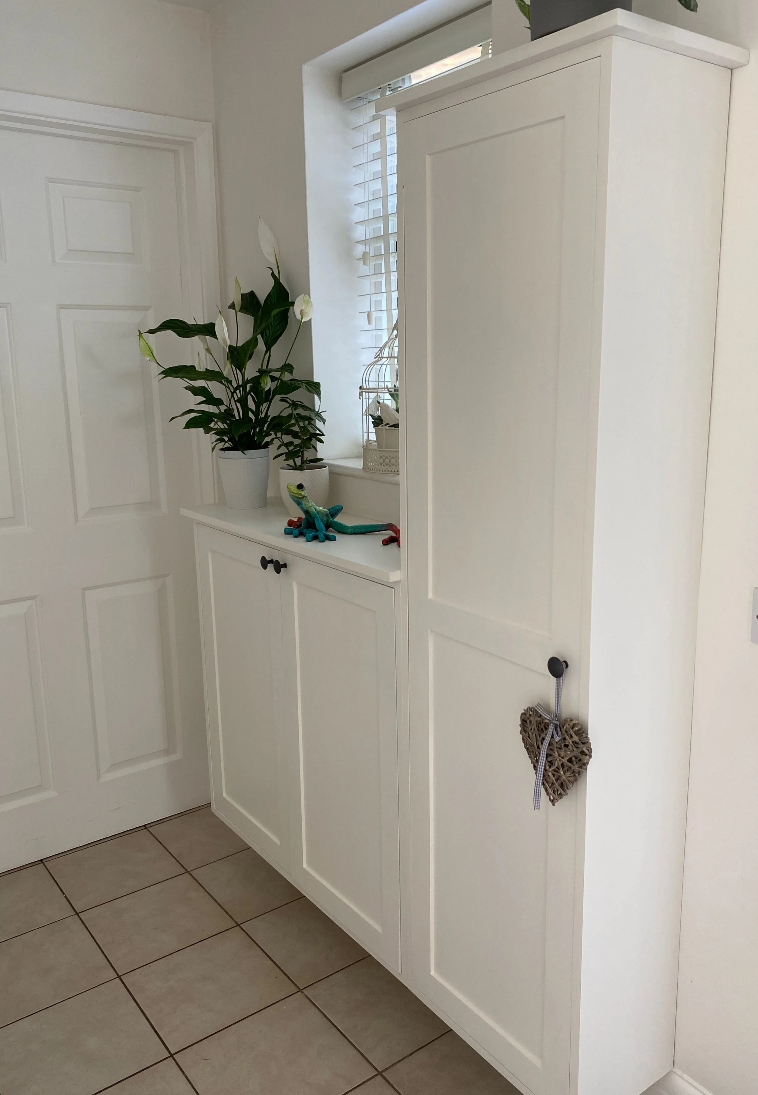 White cabinet next to a window with white blinds, decorated with a potted peace lily plant, a decorative caged plant, a toy lizard, and a heart-shaped woven ornament hanging on the door handle.