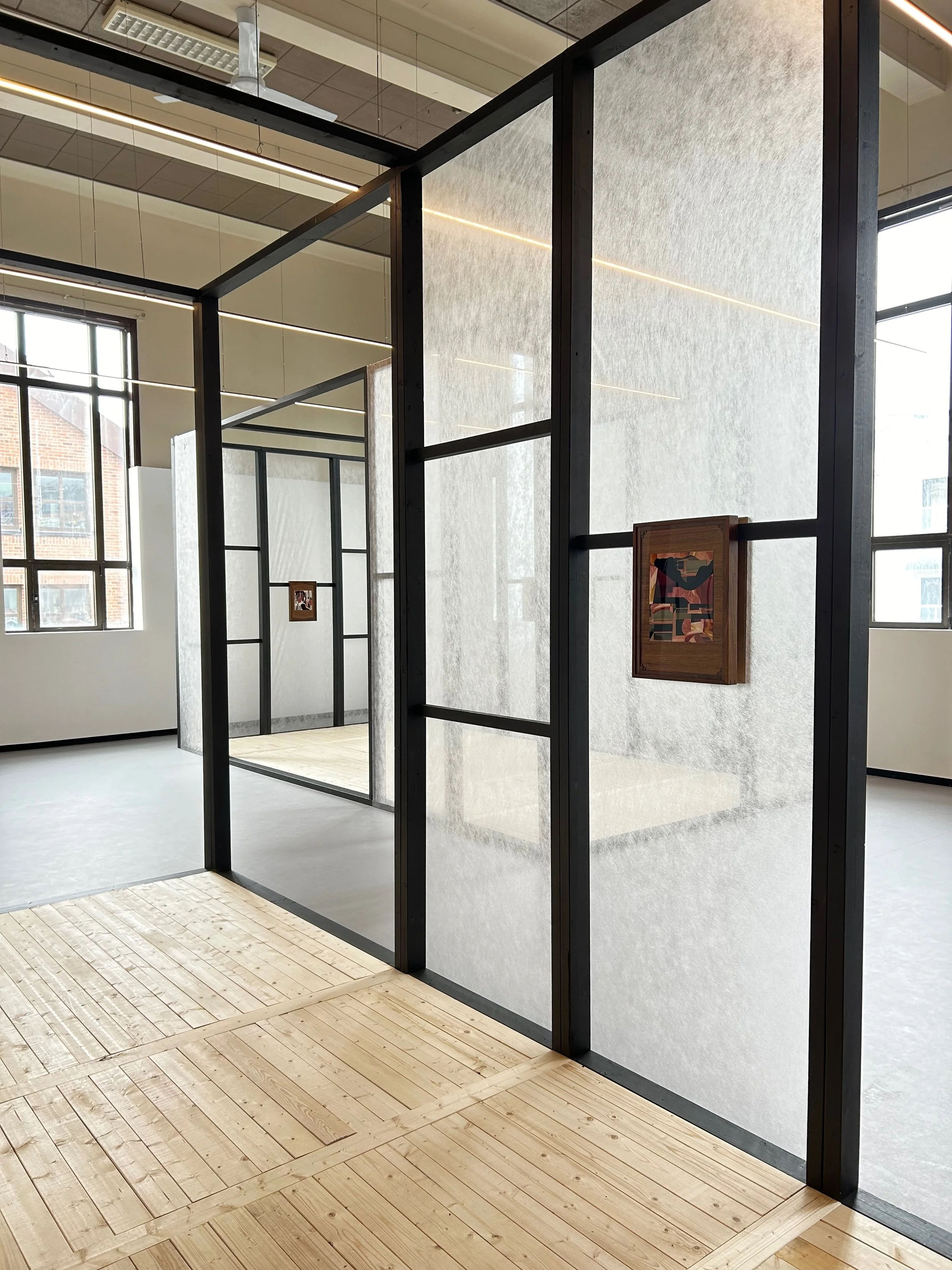 Art gallery with light-colored wooden flooring and glass and metal partitions, displaying small framed artwork on white walls, large windows allowing natural light, and a view of neighboring brick buildings.